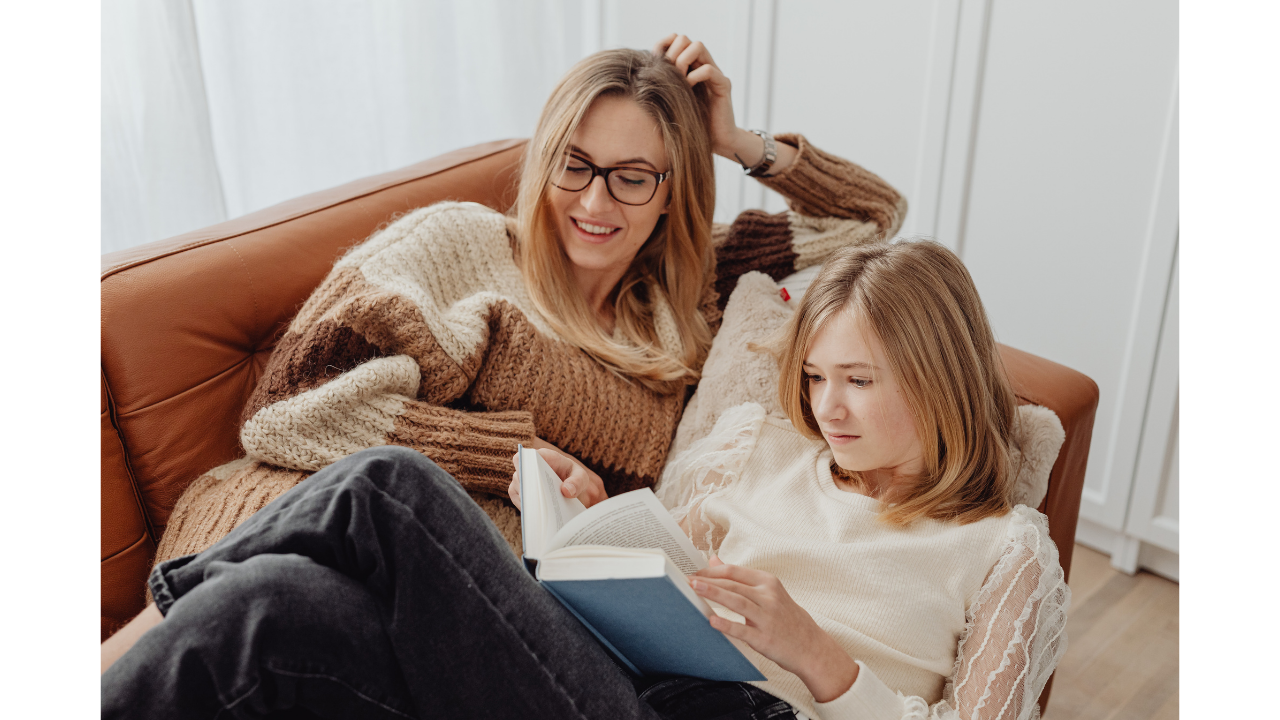 Teen girl reading a book on a couch next to her mother.