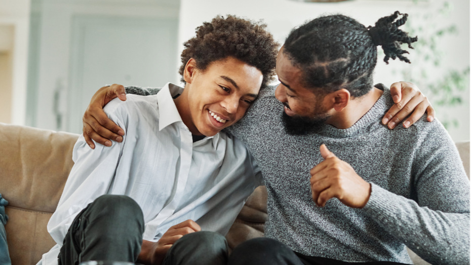 Teen boy sitting on a couch with his father's arm around him.