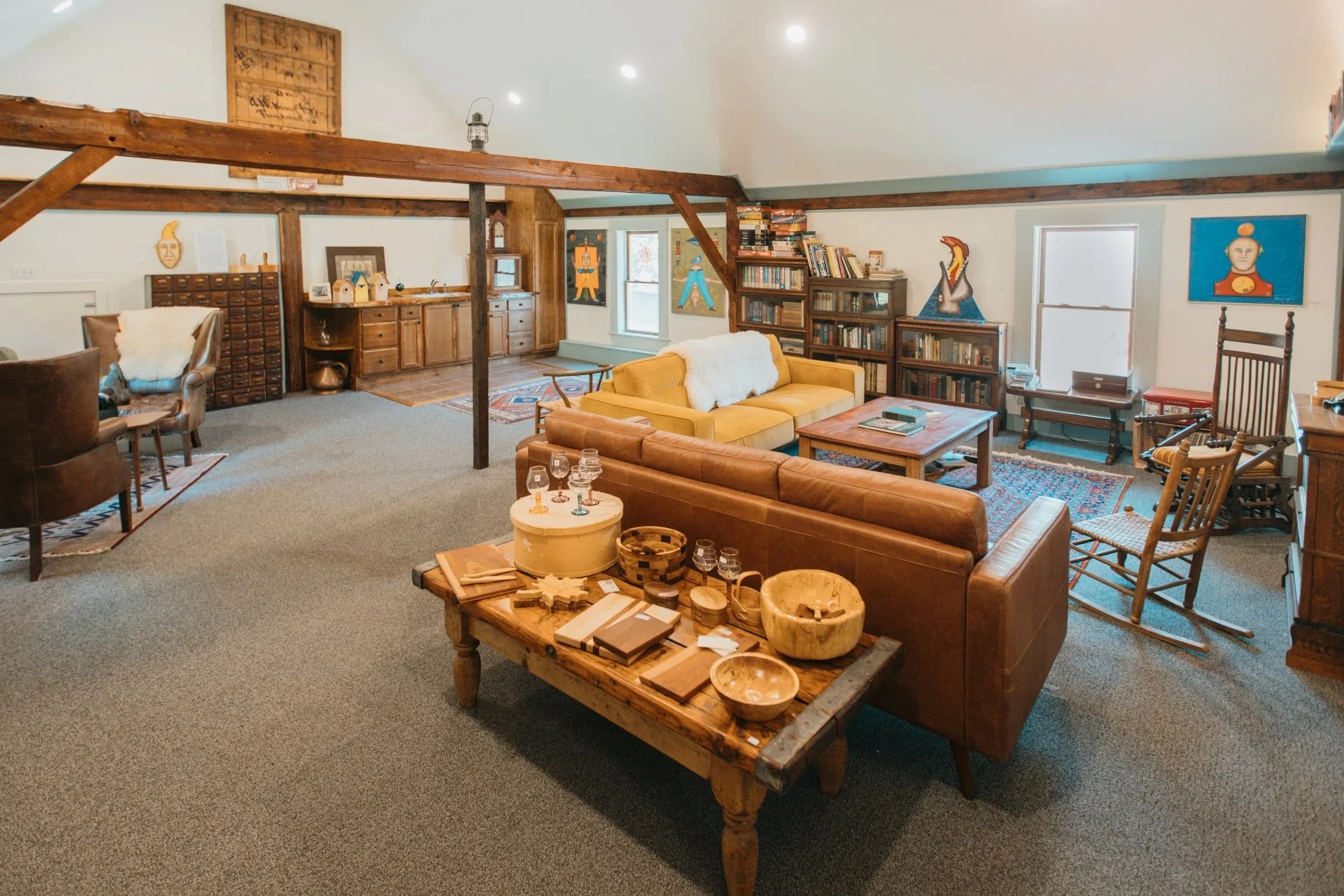 Spacious living room with vintage furniture, bookshelves, and artwork on the walls. Wooden beams and a carpeted floor.