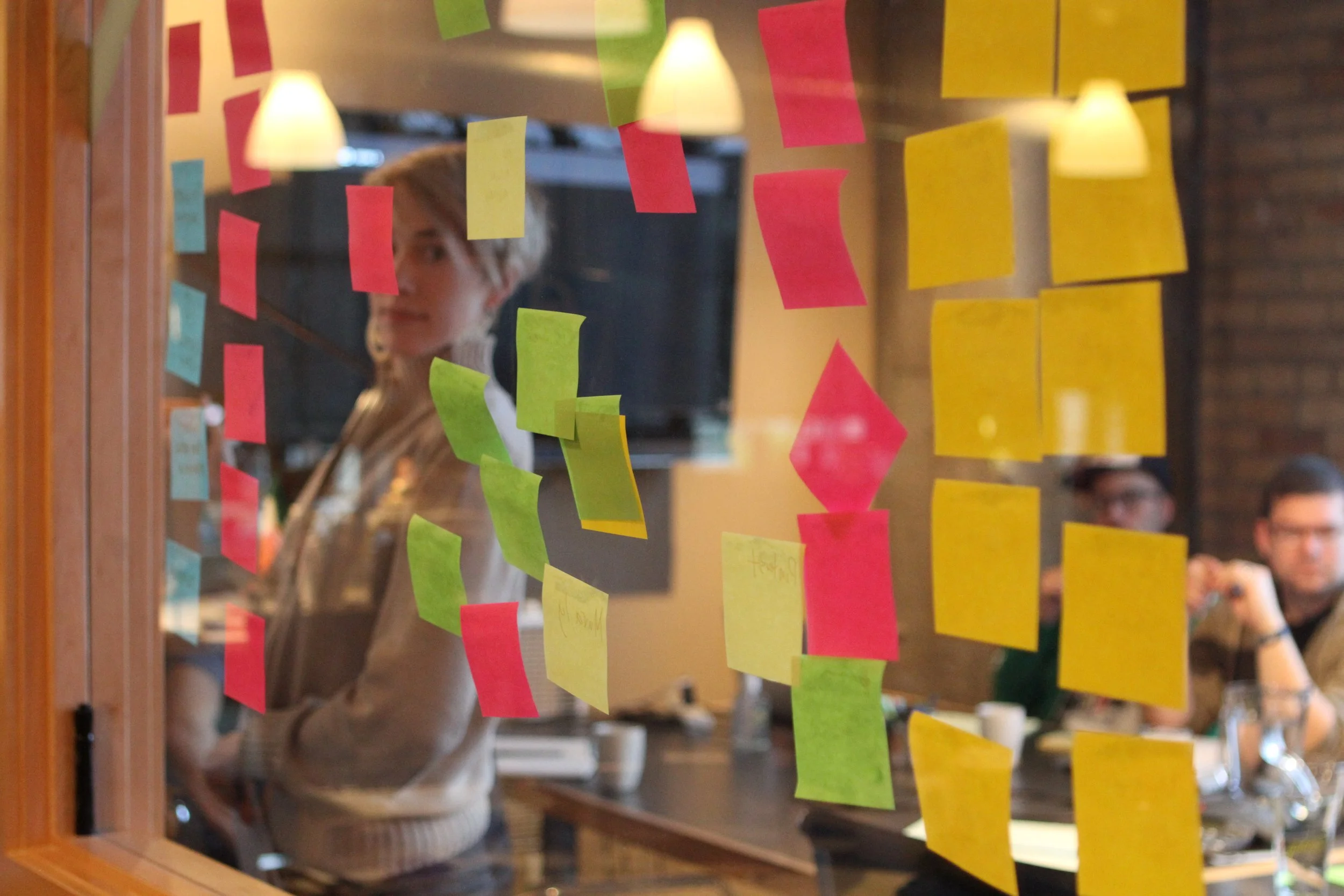 People working behind a glass window covered with colorful sticky notes in a meeting or planning session.