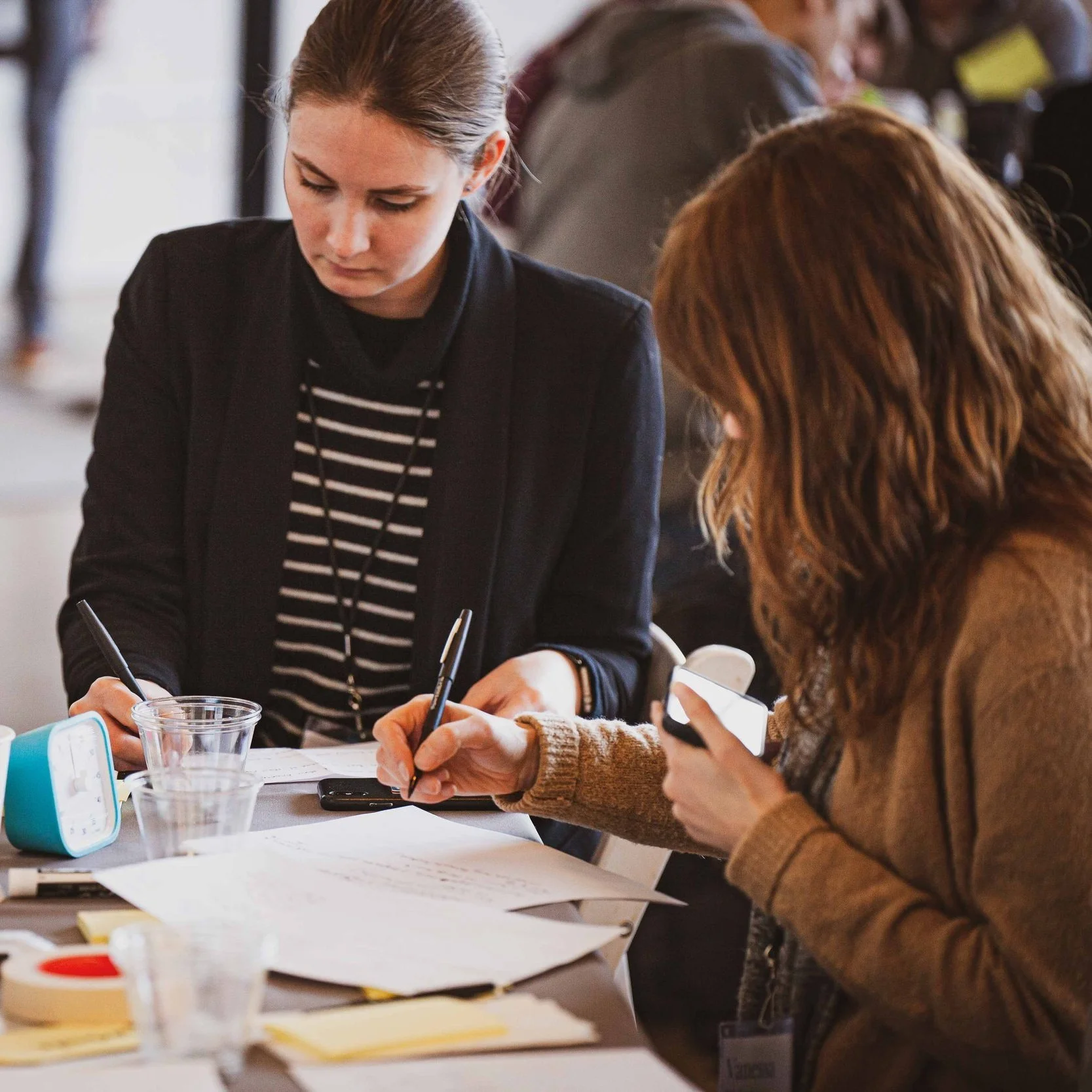Two women are sitting at a table, working with papers, pens, and a smartphone. The woman on the left has brown hair tied in a bun and is wearing a black blazer and a striped shirt. The woman on the right has wavy reddish hair and is wearing a brown jacket, looking at her phone.
