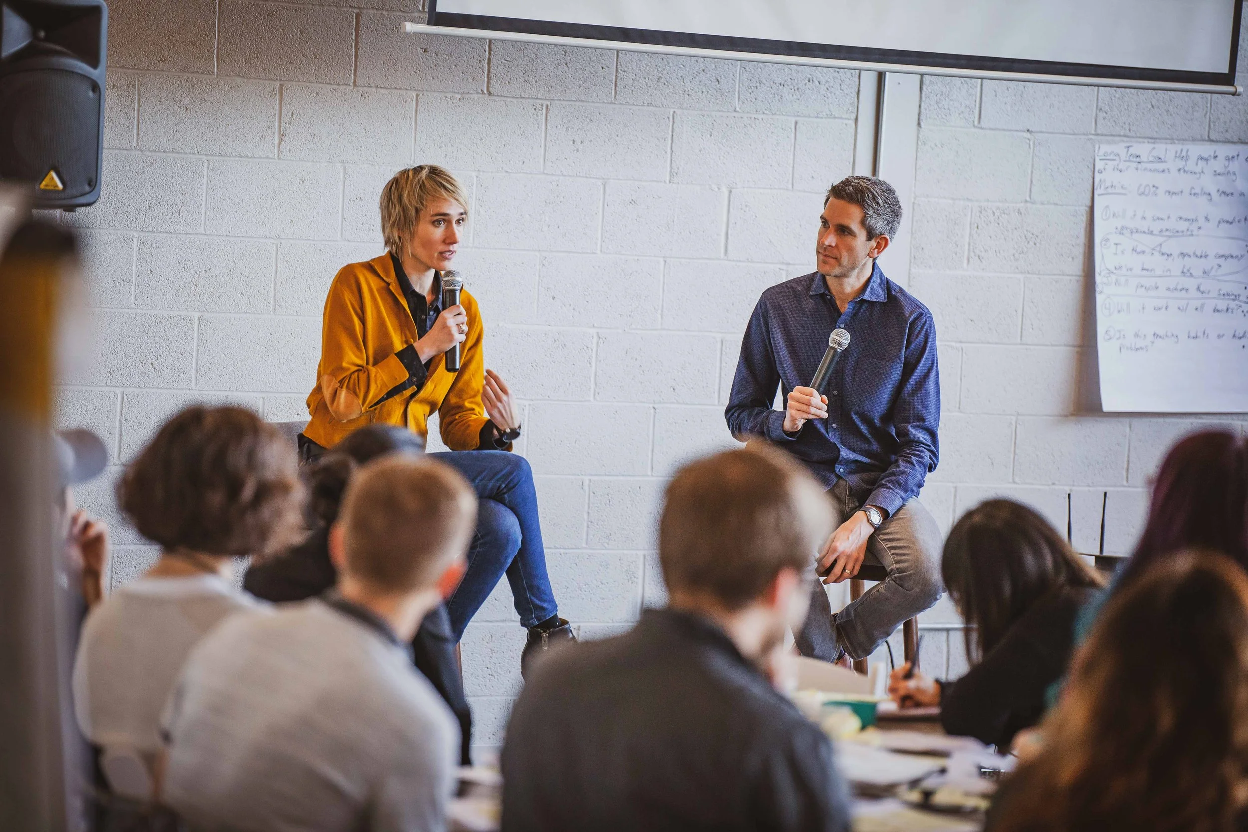 Two people with microphones speaking in front of an audience in a conference room with a white brick wall and a whiteboard.