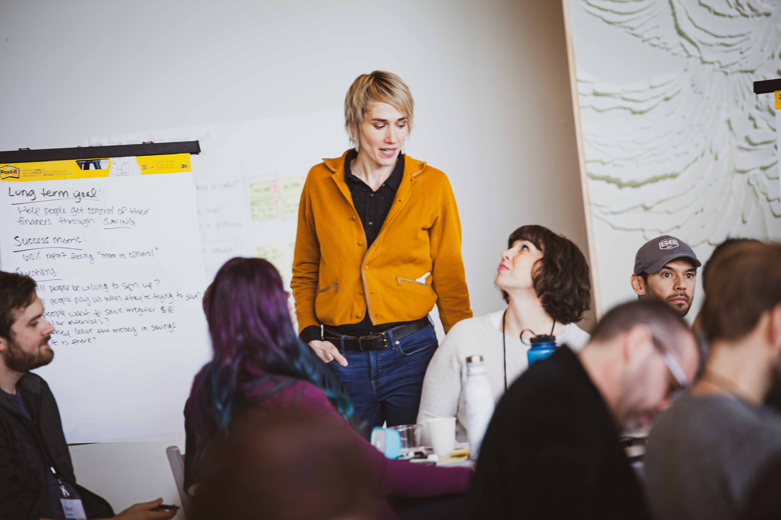 A woman in a yellow jacket standing and speaking to a seated woman with dark curly hair during a meeting or workshop, with other participants seated around a table.