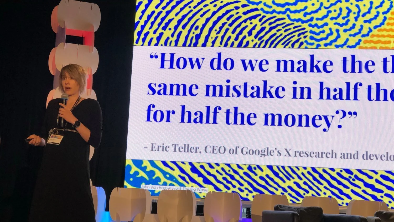 A woman with short blond hair, wearing a black dress and a lanyard, holding a microphone and speaking at a conference. Behind her is a large screen displaying a quote by Eric Teller, CEO of Google’s X research and development, that reads: 'How do we make the same mistake in half the time for half the money?' The background includes abstract yellow and blue patterns and a black backdrop.