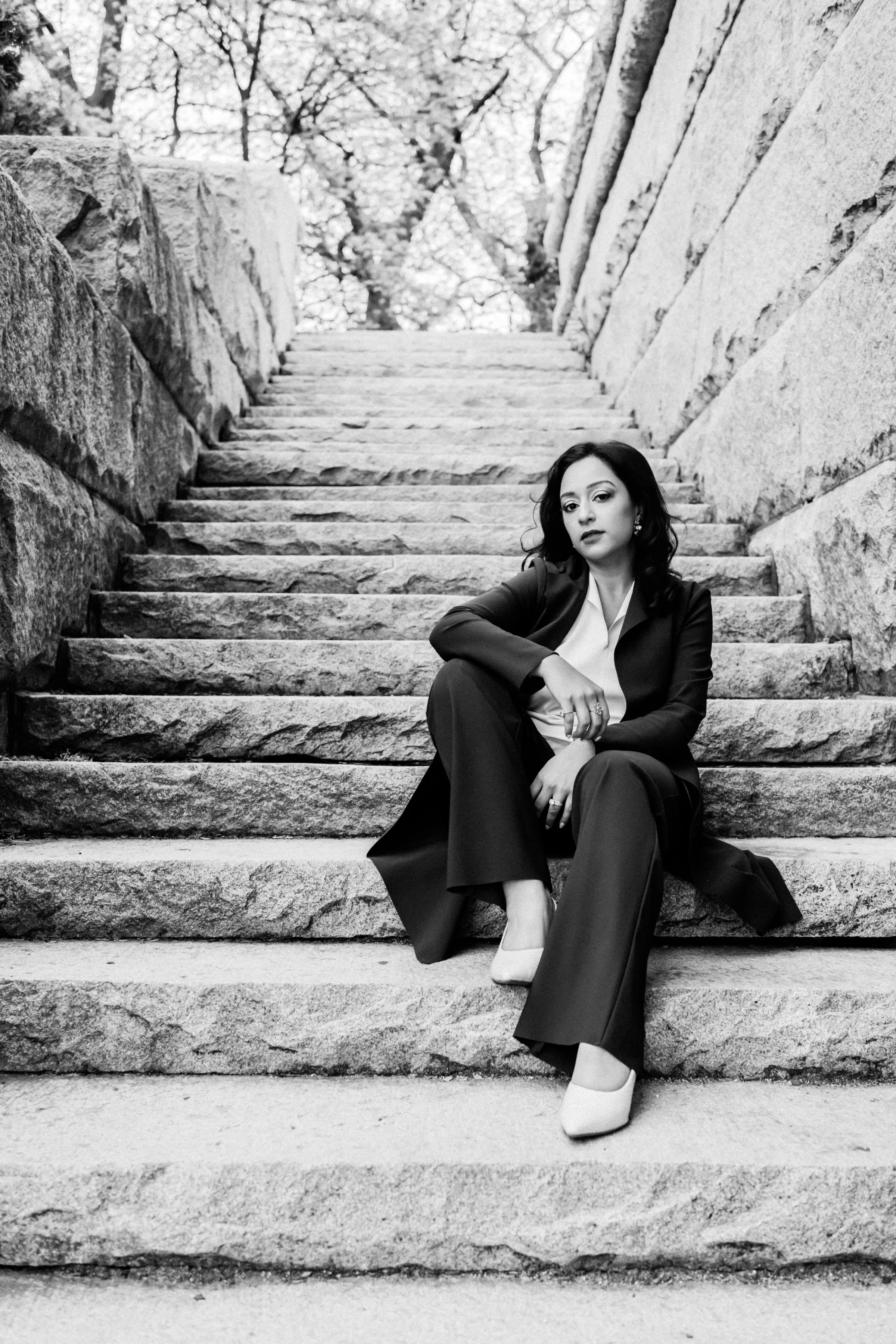 A woman sitting on stone steps outdoors, dressed in a black suit with a white blouse and beige heels, with a background of trees and stone walls.