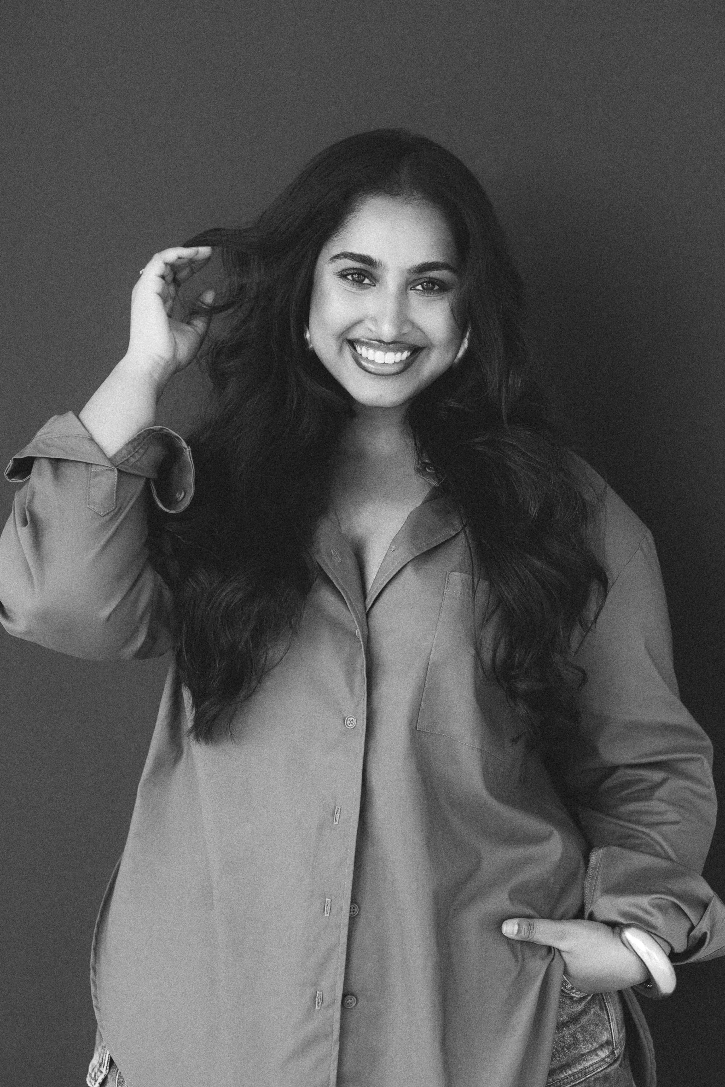 Black and white photo of a woman with long wavy hair, smiling, wearing a loose shirt, with her left hand on her hip and her right hand touching her hair.