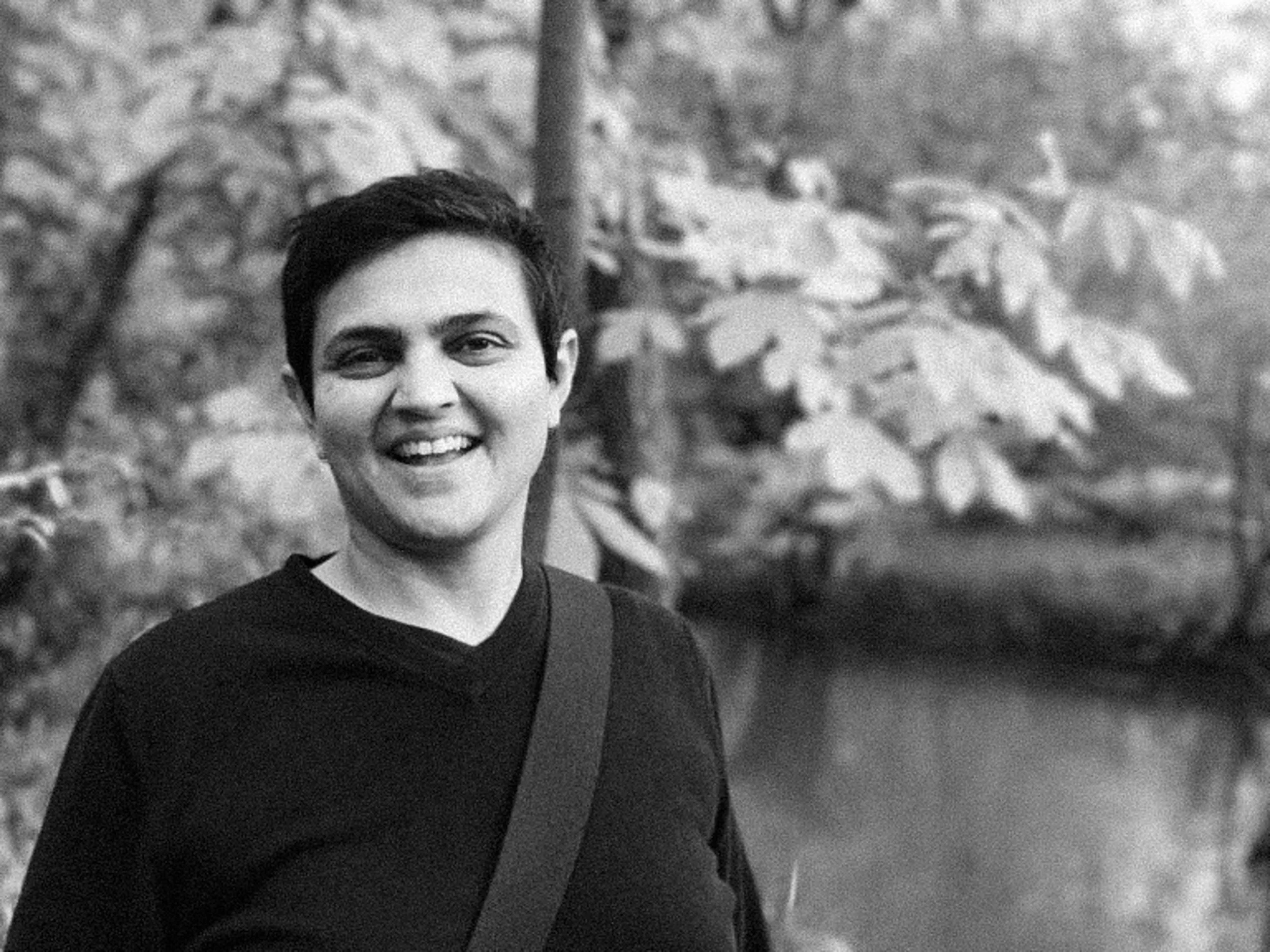 A black-and-white photo of a smiling person with short dark hair, wearing a dark shirt, standing outdoors with trees and foliage in the background.