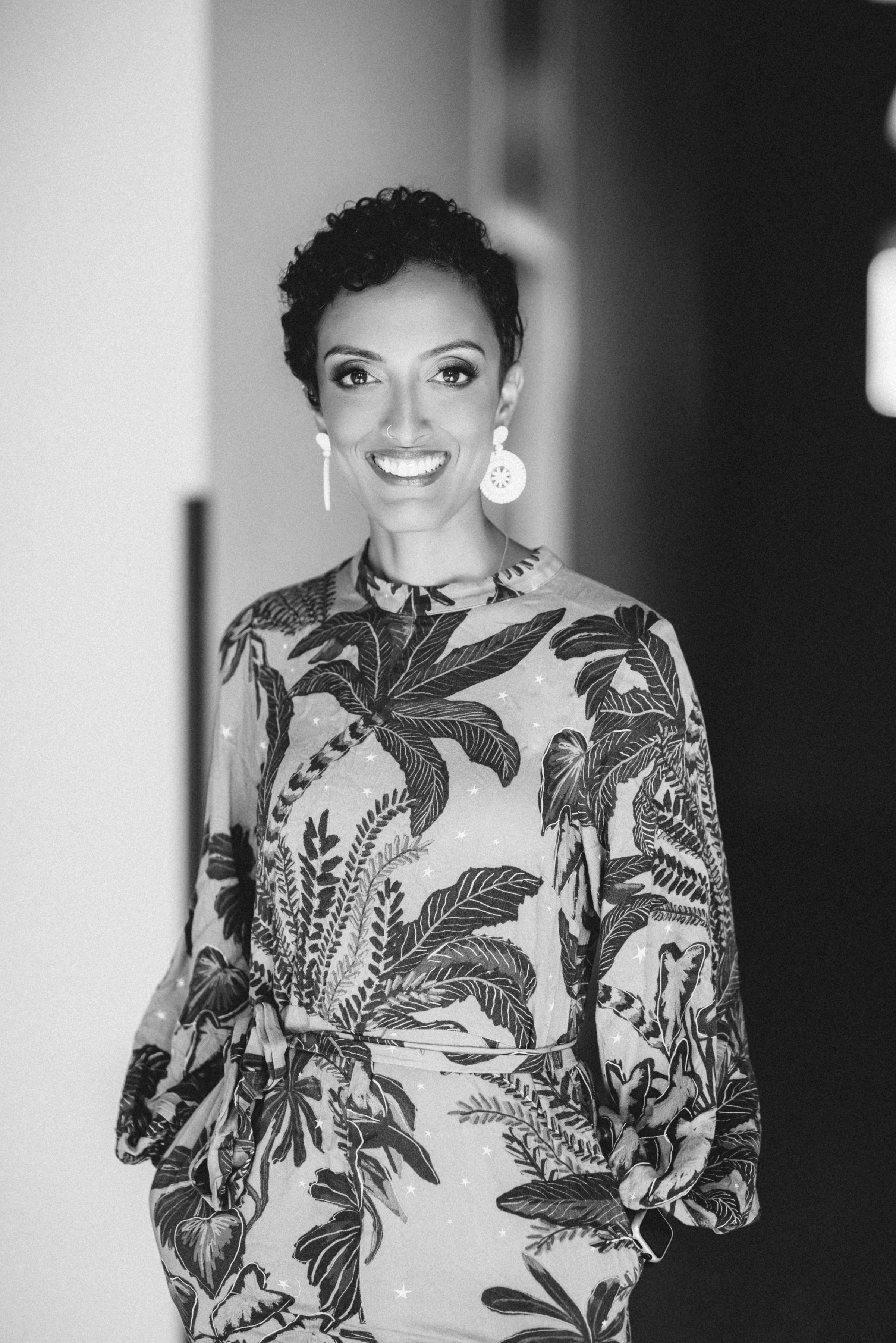 Black and white photograph of a woman with short curly hair, wearing hoop earrings and a tropical print dress, smiling at the camera.