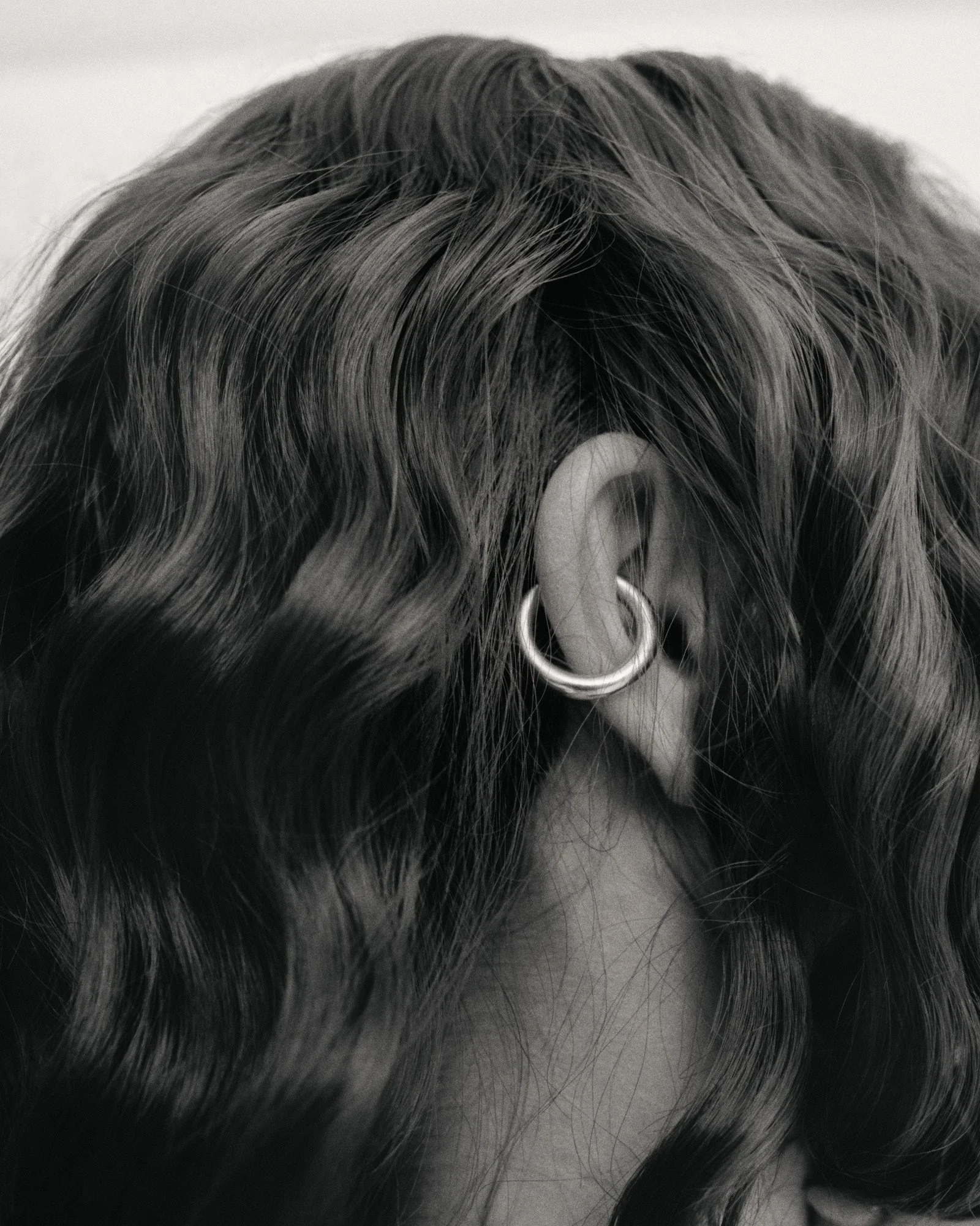 Close-up black and white photo of a person's head showing textured, wavy hair and an ear with a hoop earring.