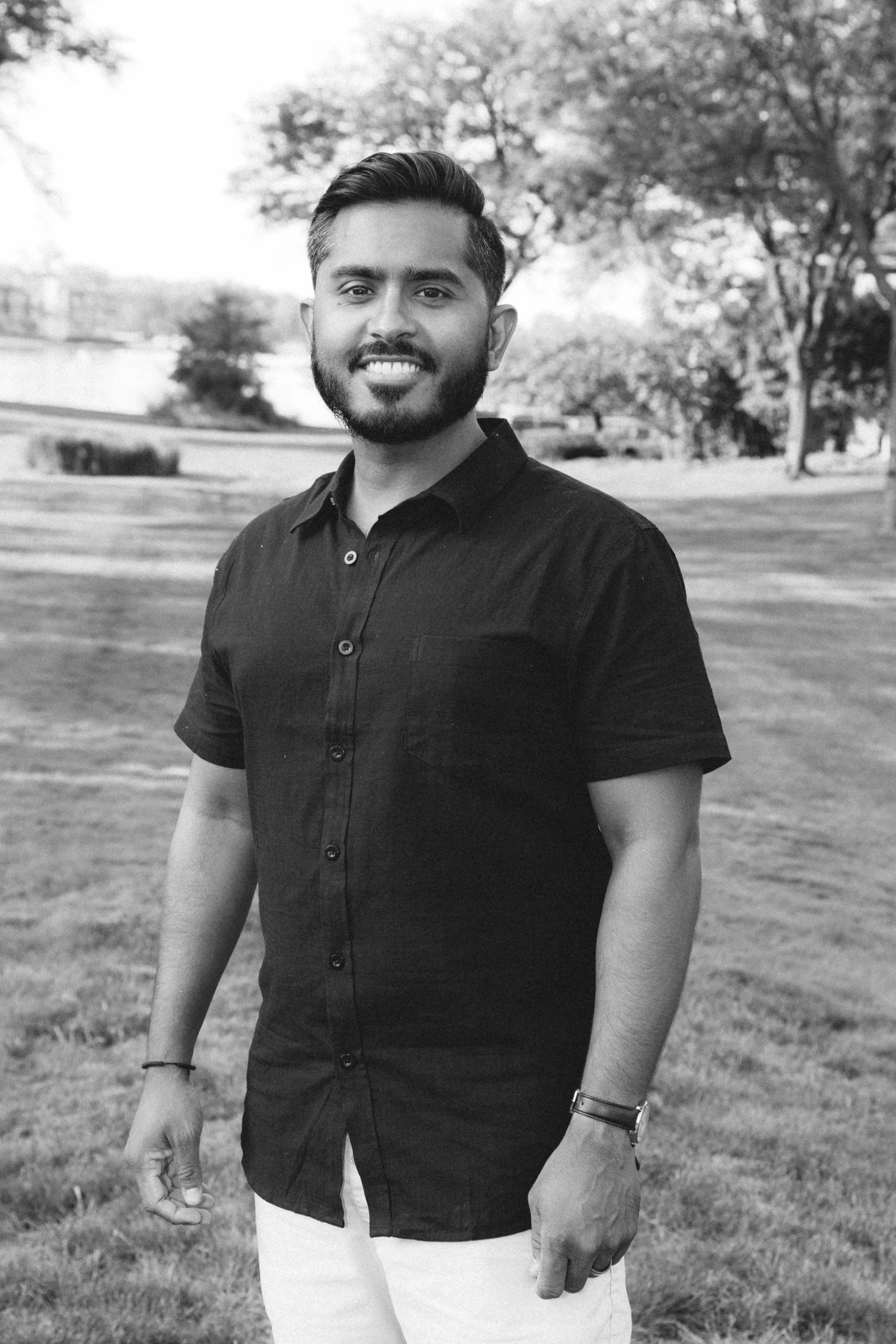 A young man with a beard and styled hair smiling outdoors in a park setting, wearing a dark short-sleeved button-up shirt and a watch, with trees and grass in the background.