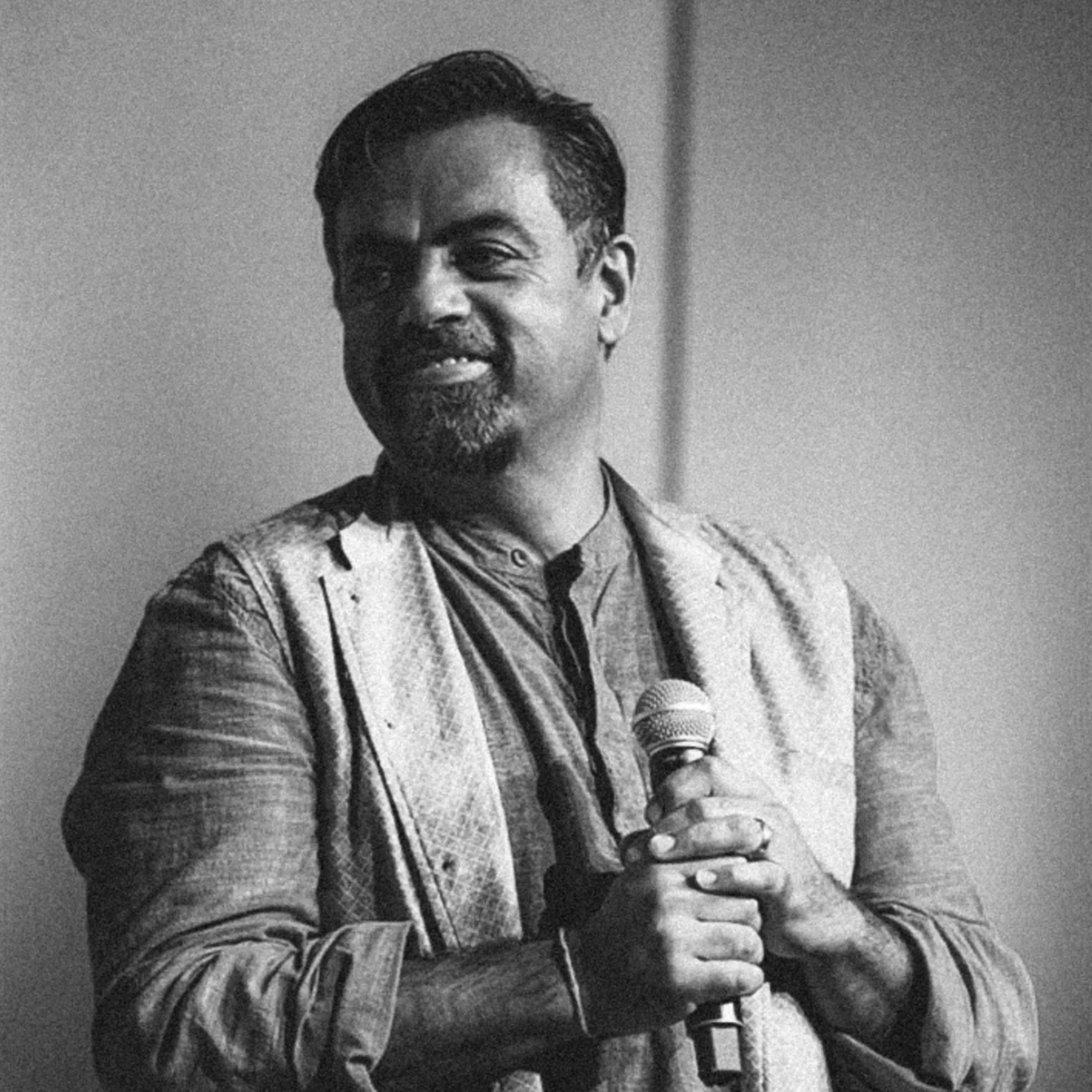 A black and white photo of a man smiling while standing behind a microphone, wearing a patterned shirt.