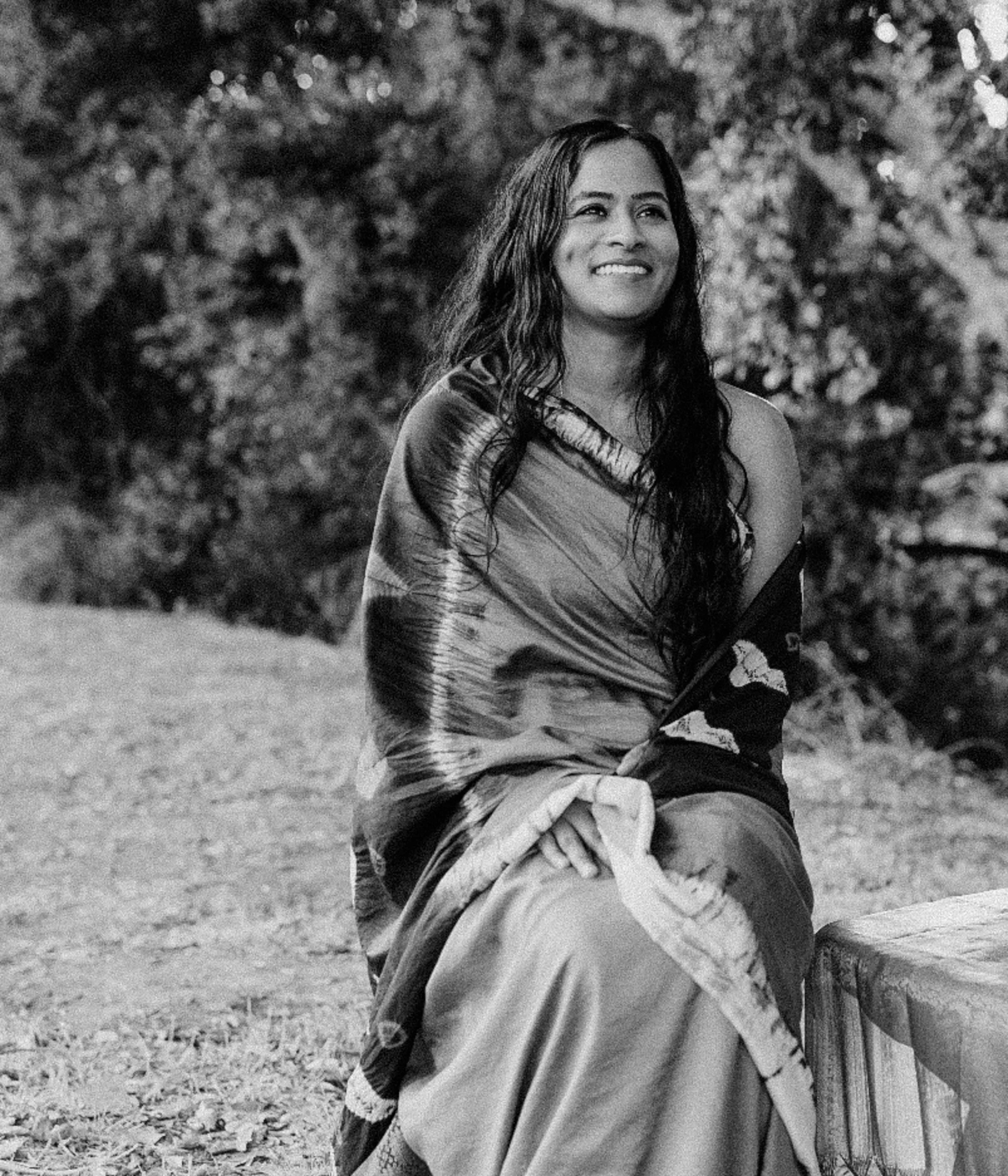 A woman sitting outdoors on grass, wearing traditional attire and smiling, with trees in the background.