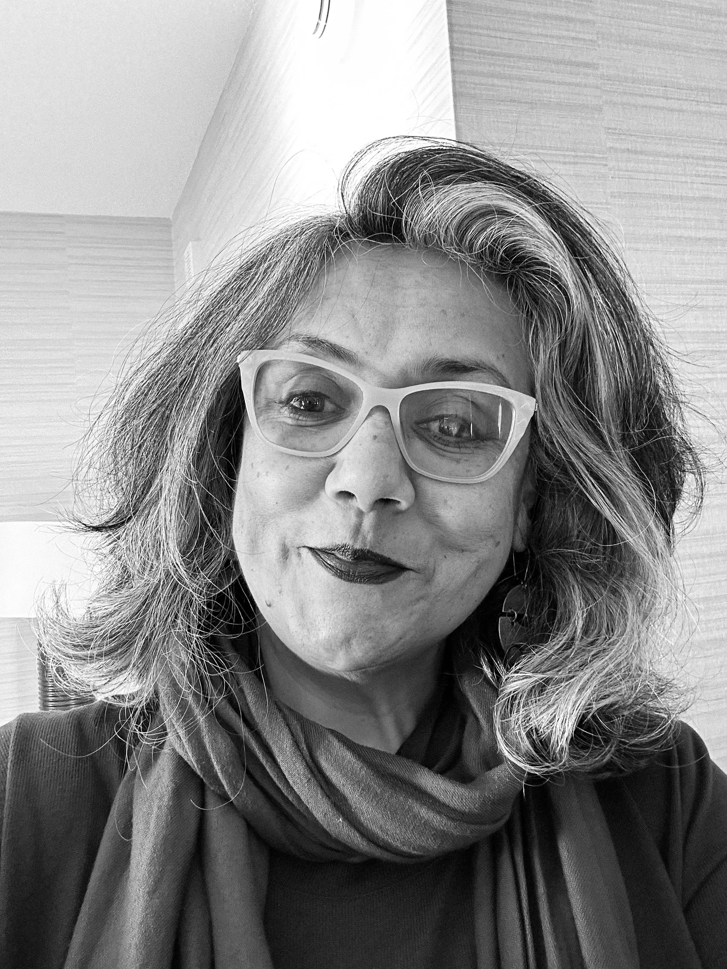 Close-up of a woman with curly hair and glasses, wearing a scarf and jacket, taking a selfie indoors.