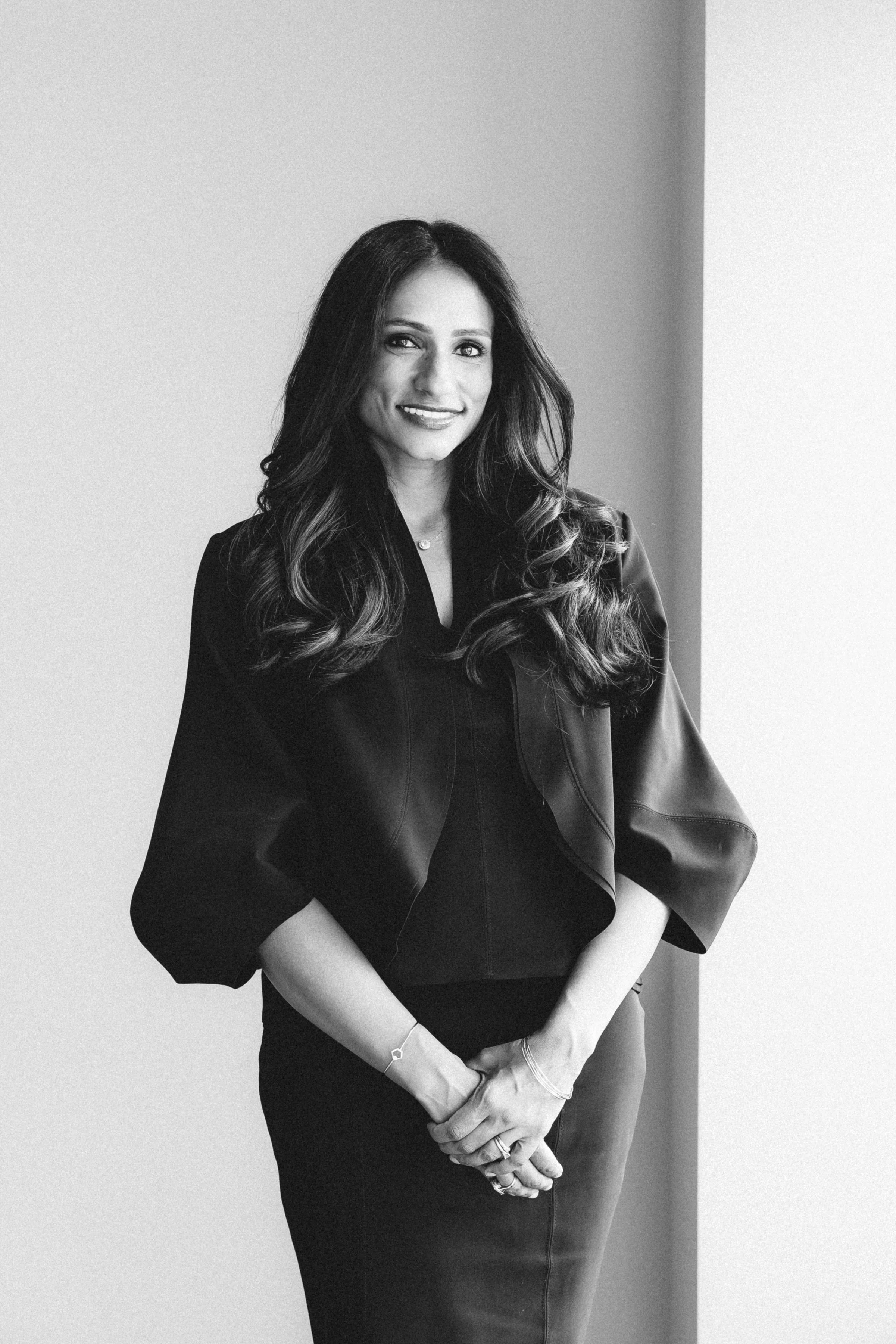 Black and white photo of a woman with long wavy hair, smiling, dressed in a dark top with puffed sleeves, standing against a plain background.