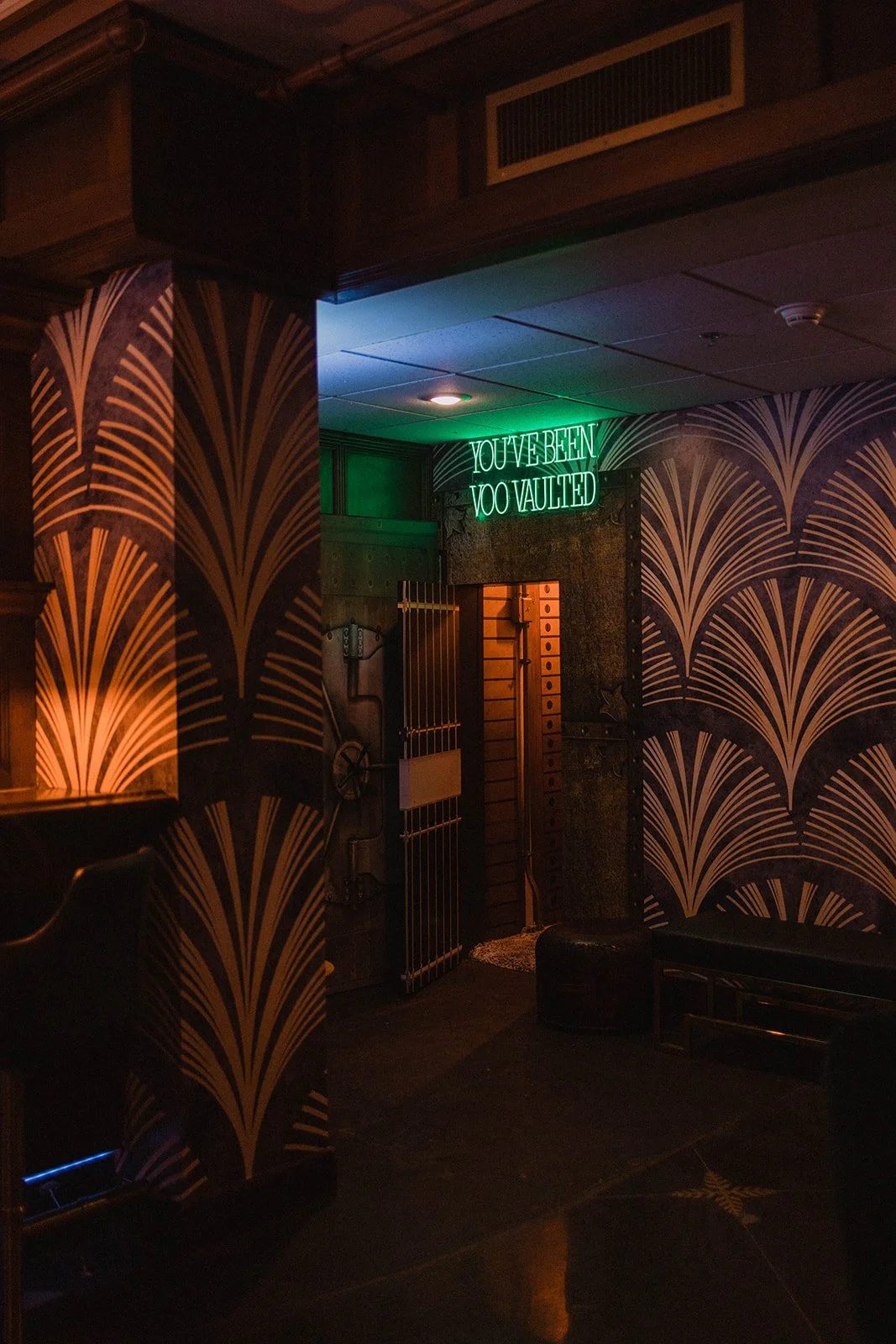 A dimly lit interior space with art deco style wallpaper, a green neon sign that reads 'YOU'VE BEEN TOO VAULTED', and a partially open door revealing wooden drawers inside a vault.