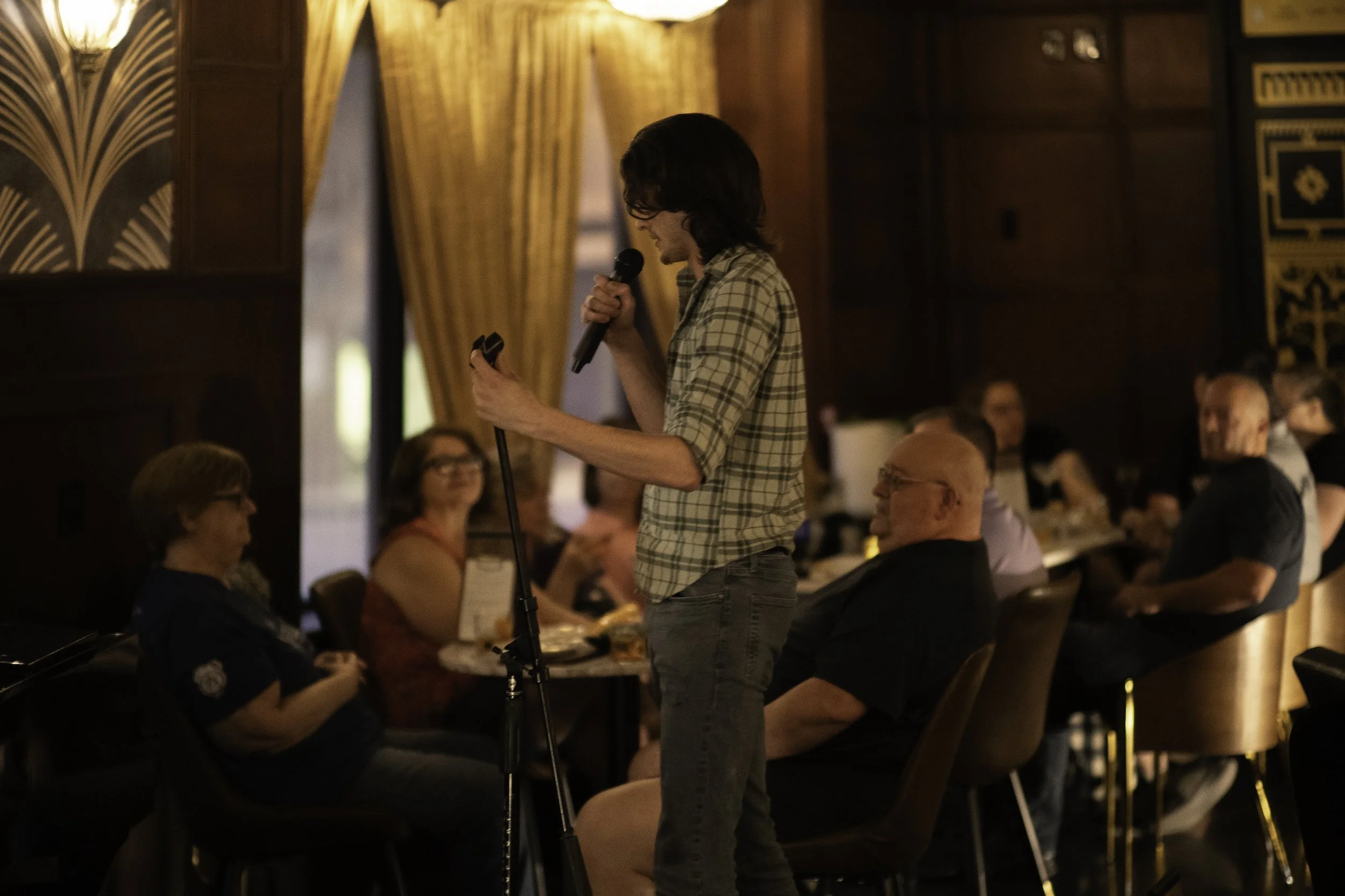 A man holding a microphone and looking at his phone in front of an audience seated at tables in a dimly lit room with golden curtains.