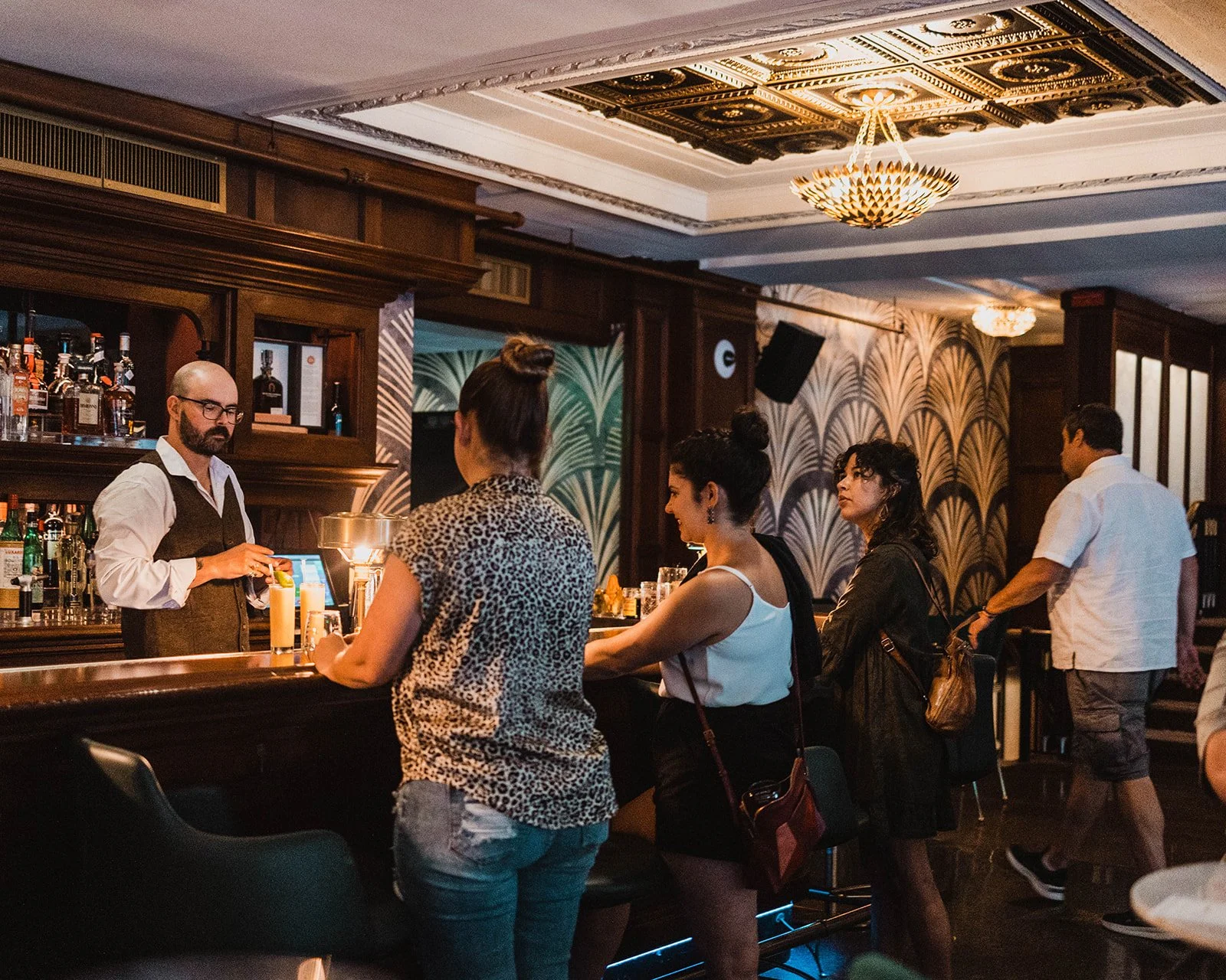 People standing in line at a bar, being served by a bartender in a warmly lit, elegant interior with vintage-style decor and patterned wallpaper.