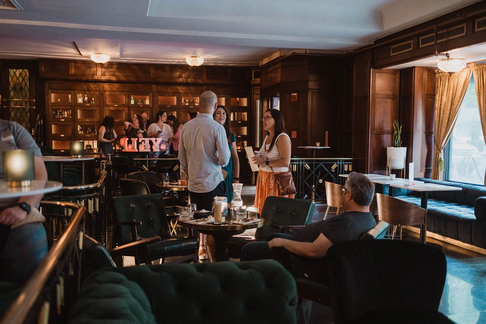 People socializing in a dimly lit restaurant or bar with wooden walls, tables, and chairs, some with drinks, and a large window with curtains on the right.