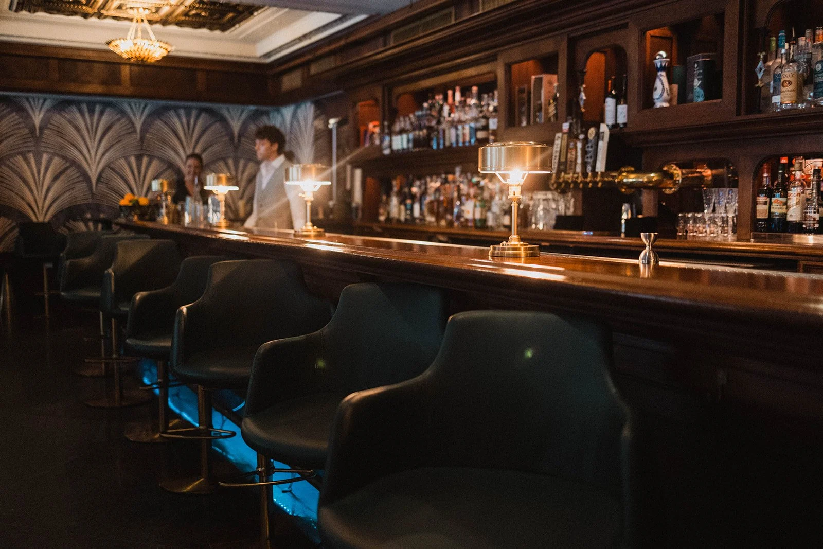 Interior of a dimly lit bar with a long wooden counter, bar stools, and bottles of alcohol on wooden shelves in the background. Two bartenders are present in the background.