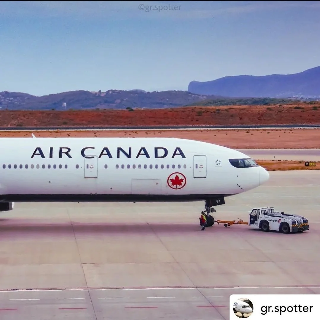 An Air Canada 777-300ER parked at the airport, with a groundcrew working near the airplane's nose wheel, and a tug vehicle attached to the aircraft.
