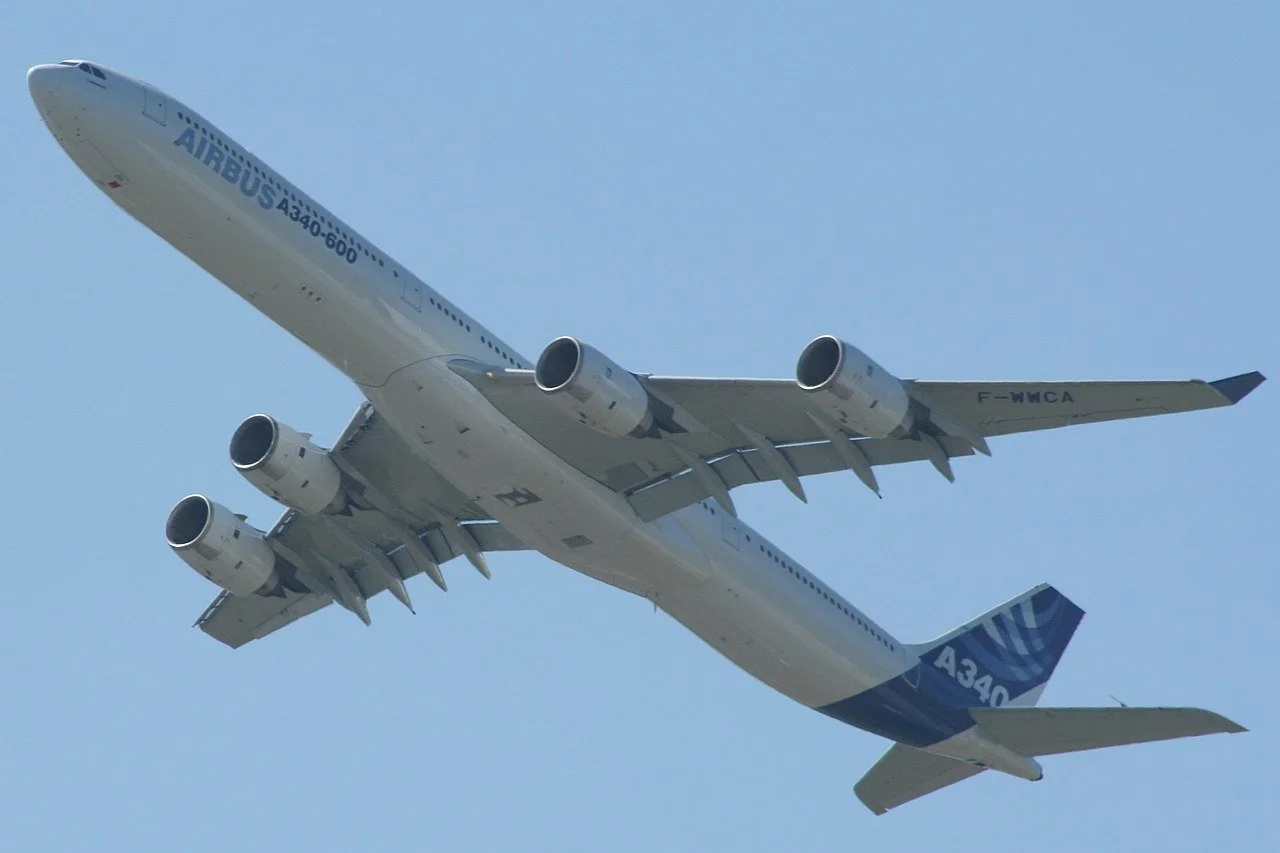A white Airbus A340 airplane flying in the sky with four engines and blue accents, seen from below.