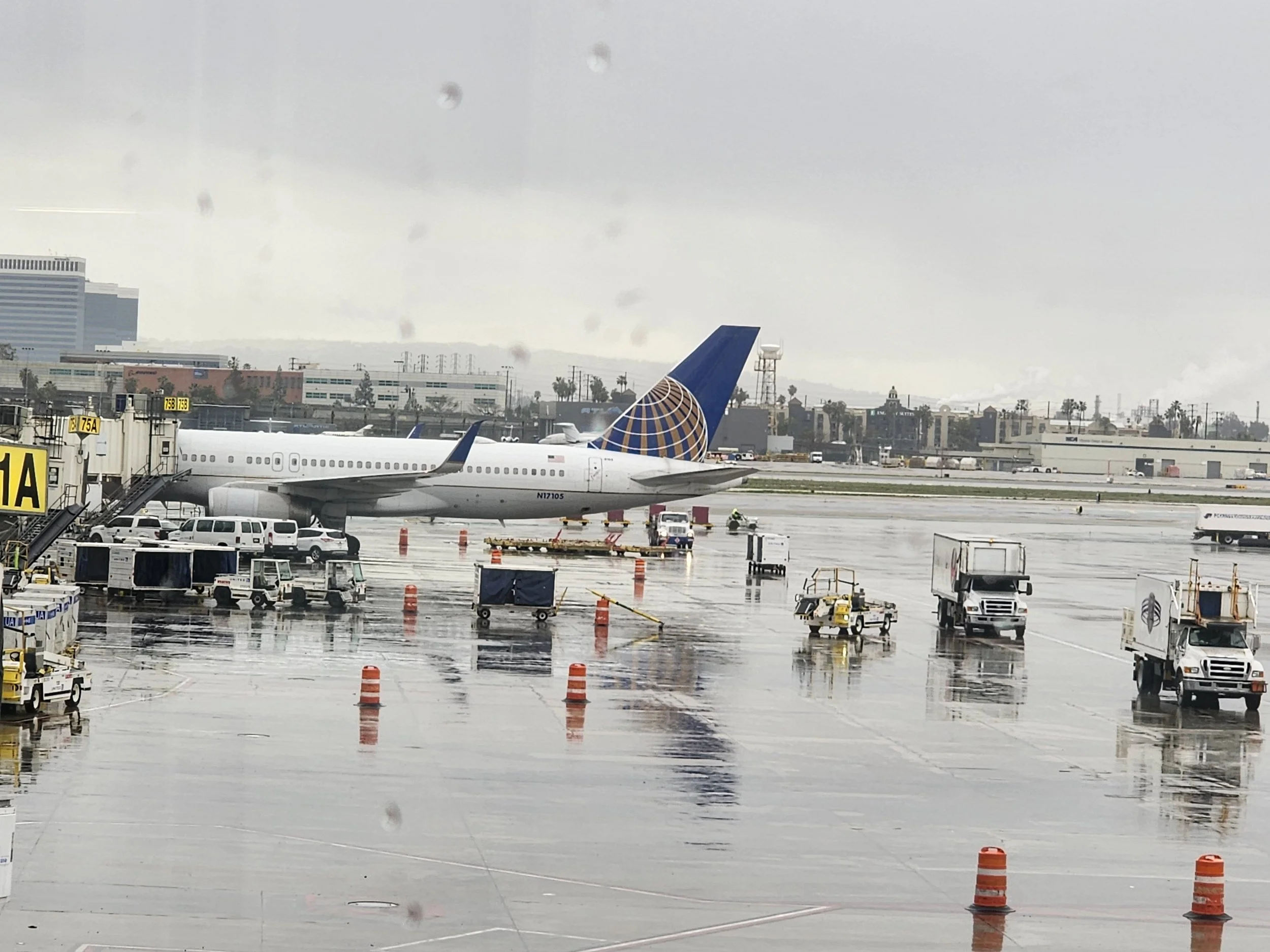 A United Airlines airplane parked at a gate on a rainy day at the LAX, with multiple ground service vehicles and wet apron