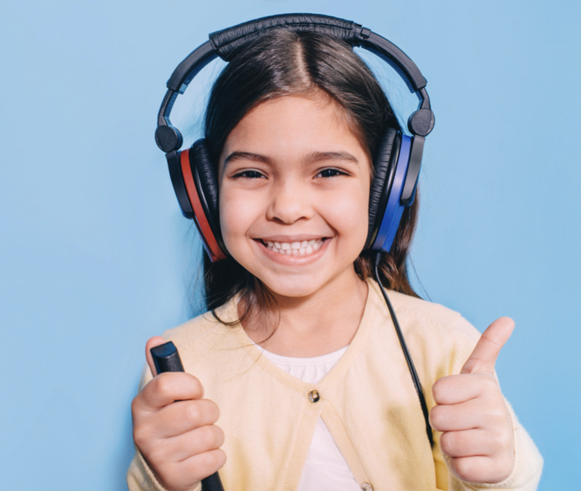 Smiling girl wearing headphones and holding a microphone, giving a thumbs up; blue background.