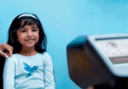 Young girl smiling, sitting in front of medical equipment, indoors with a blue background.
