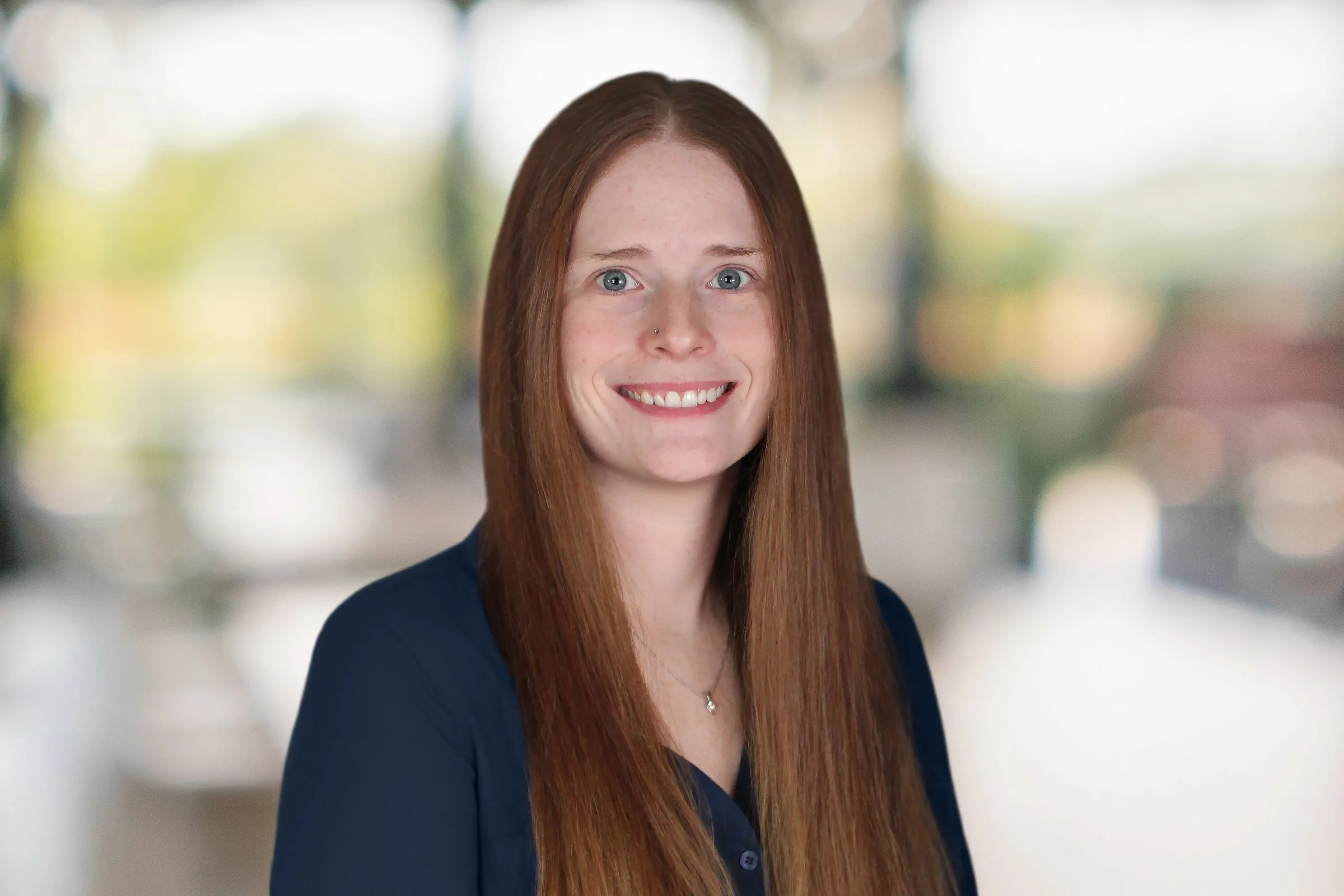 A smiling middle-aged woman with shoulder-length light brown hair, wearing a white and maroon striped shirt, standing in front of a blurred background with natural colors.