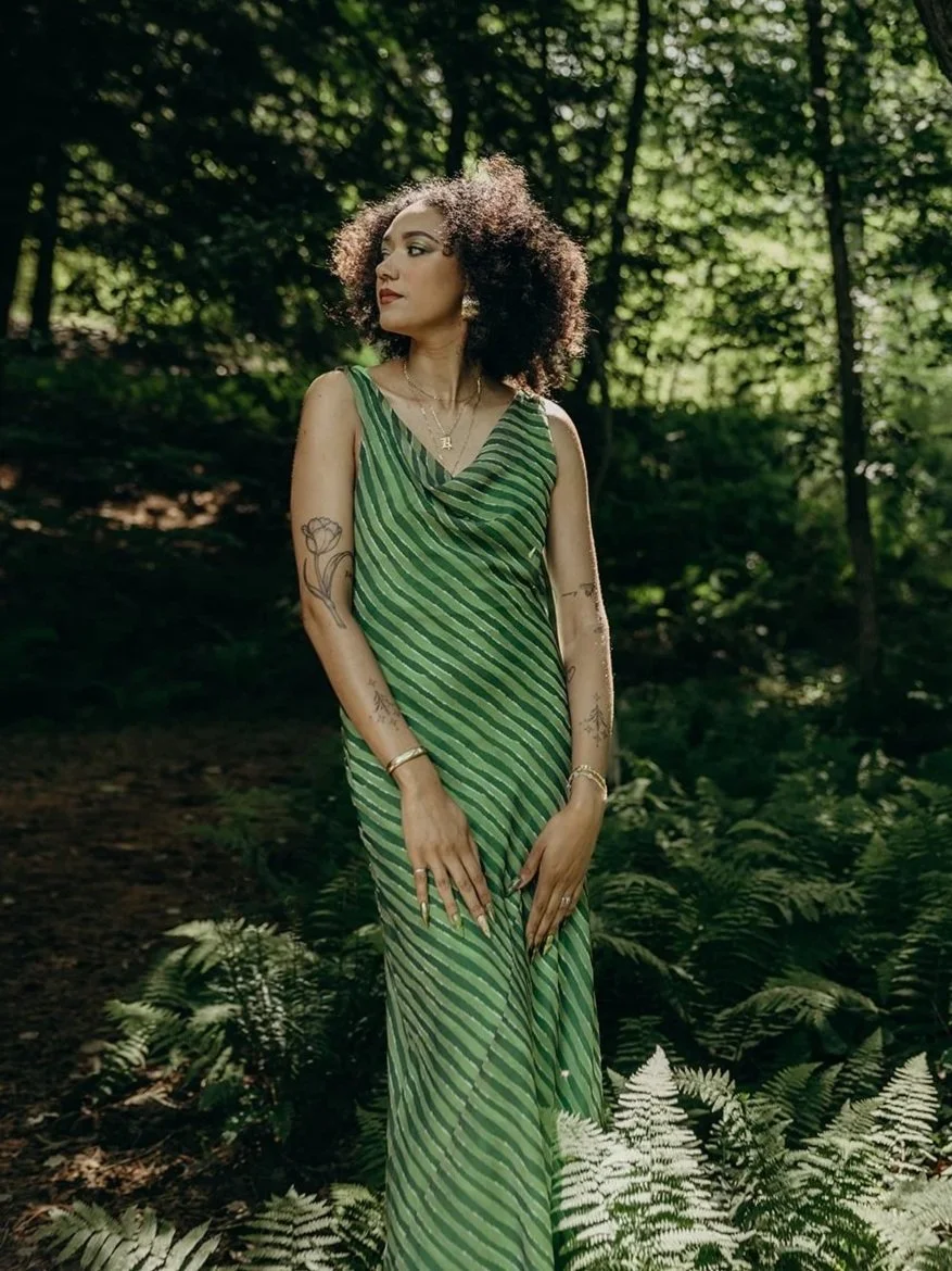 Woman standing in a forest, wearing a green striped sleeveless dress with curly hair.