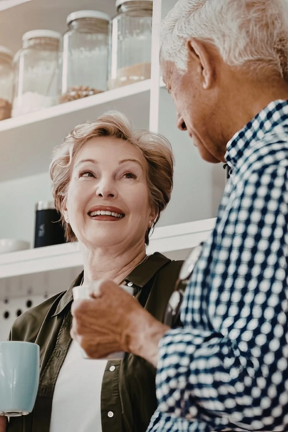 An elderly woman smiling and looking up at an elderly man in a checkered shirt, who is showing her something in his hand while holding a coffee mug. They are in a kitchen with glass jars on shelves in the background.