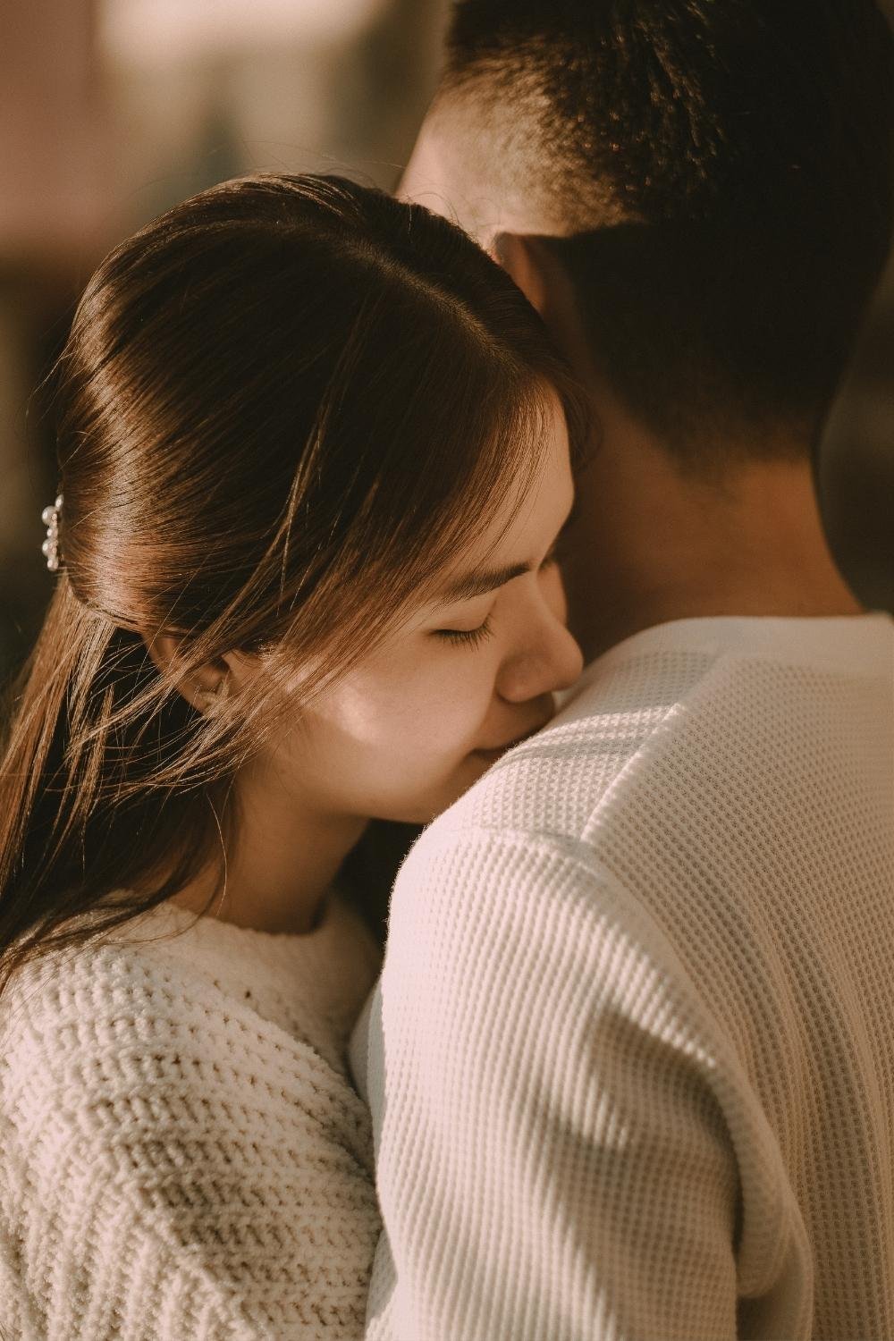 Close-up of a woman with closed eyes and long brown hair, hugging a man with short black hair, both facing each other with probably their foreheads touching