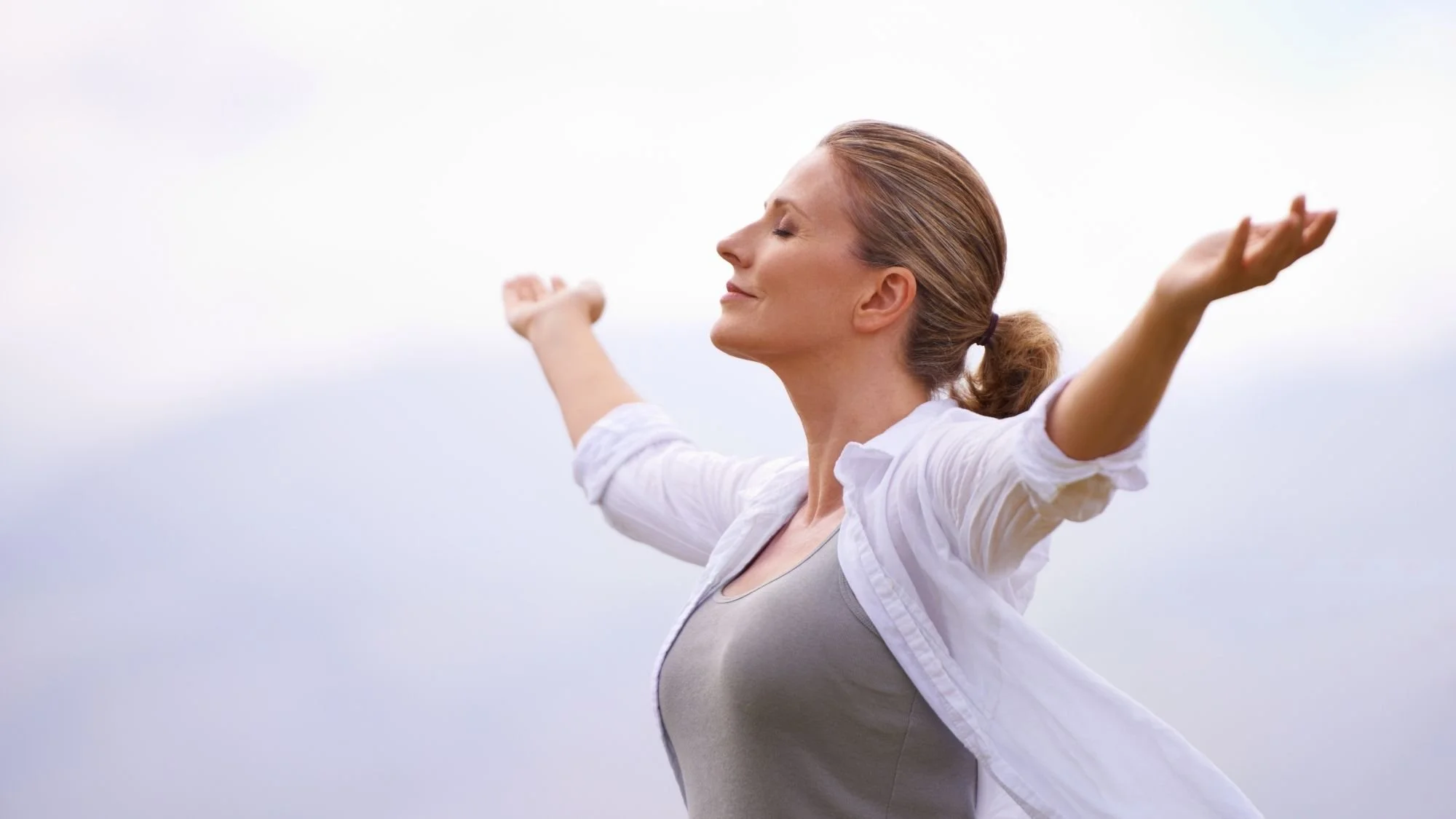 Woman outdoors with arms outstretched, eyes closed, enjoying the breeze on a cloudy day.