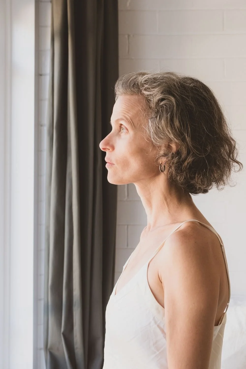 A woman with wavy gray hair looking out a window with dark curtains.