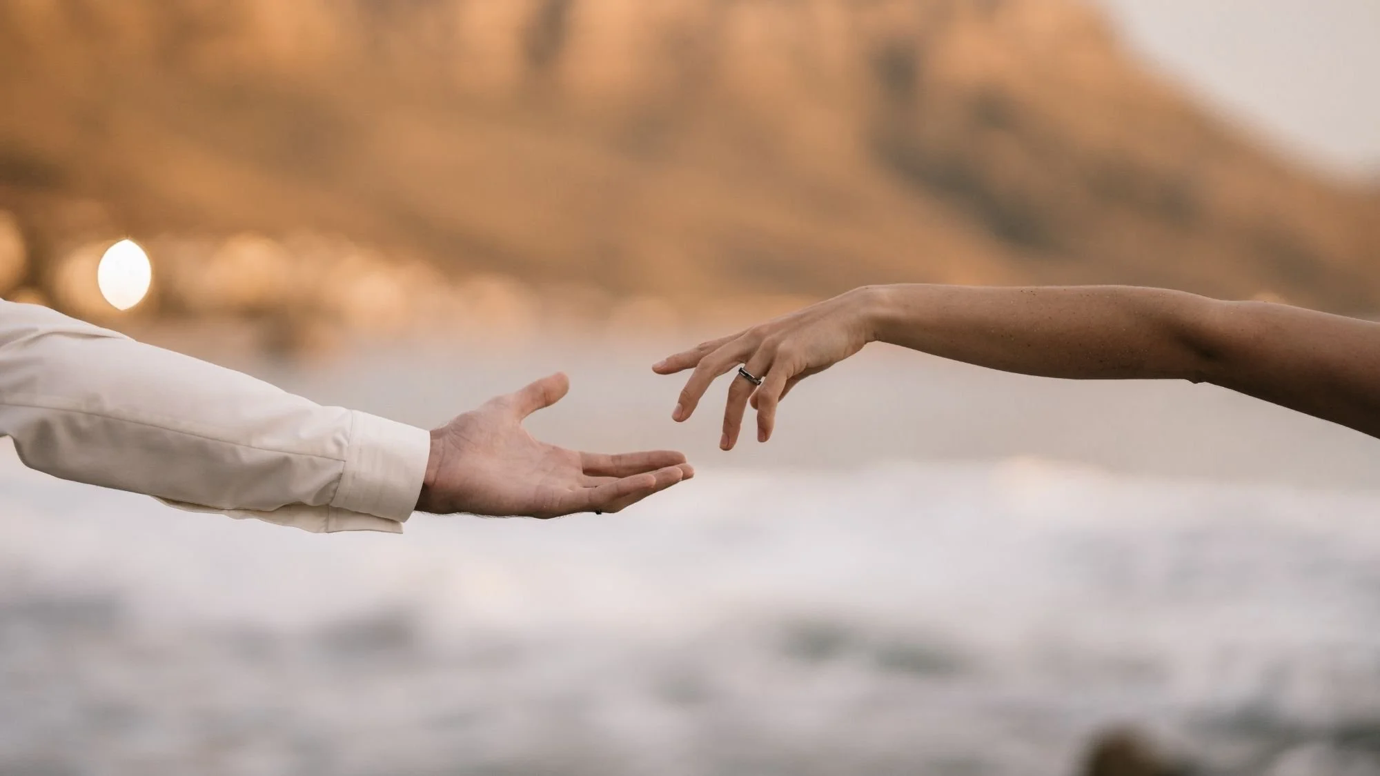 A person reaching out their hand to another person, with both hands extending from opposite sides of the image. The background is blurred with warm colors.