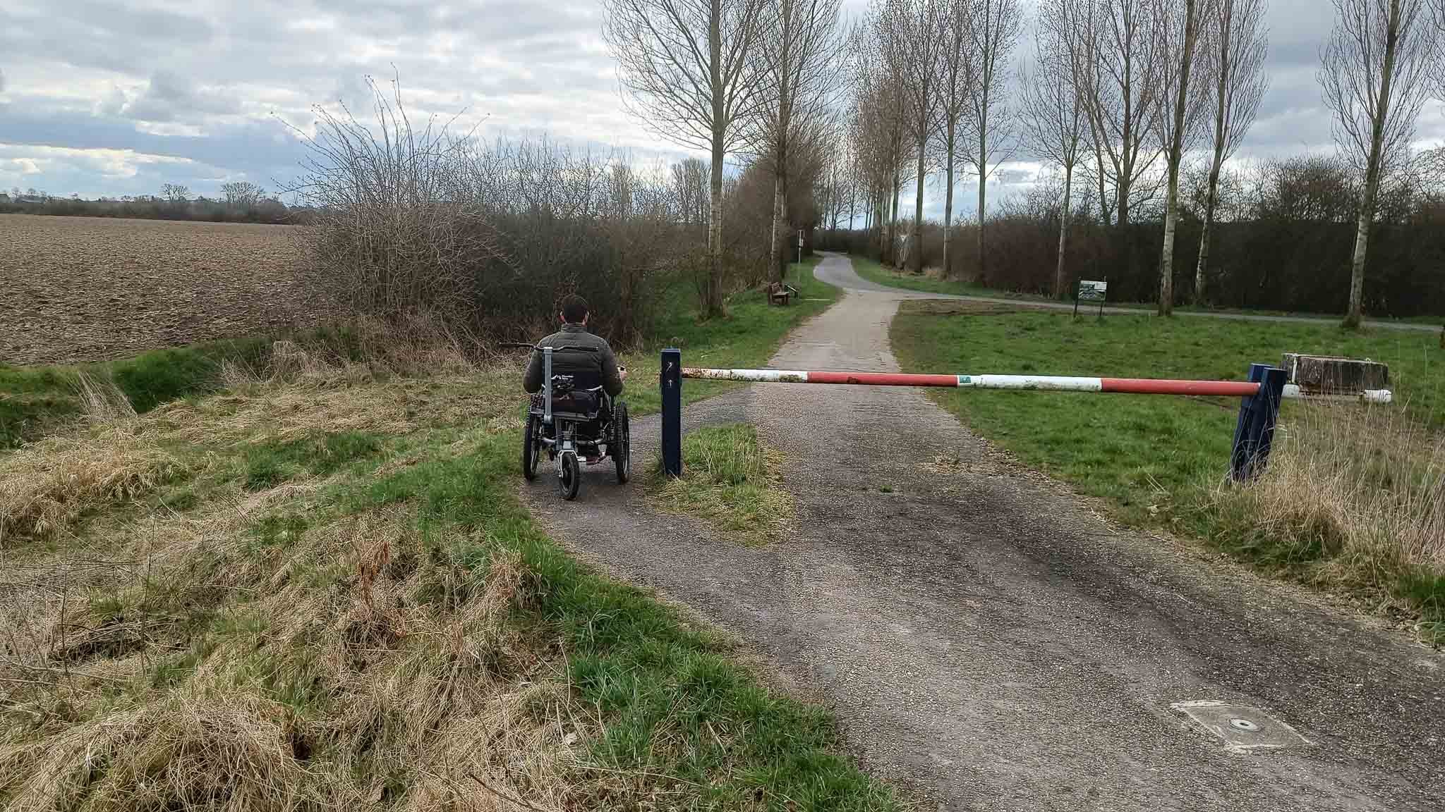 Man in een rolstoel voor een laag hek op een zandweg in een landelijk gebied met boomgaarden en grasvelden.