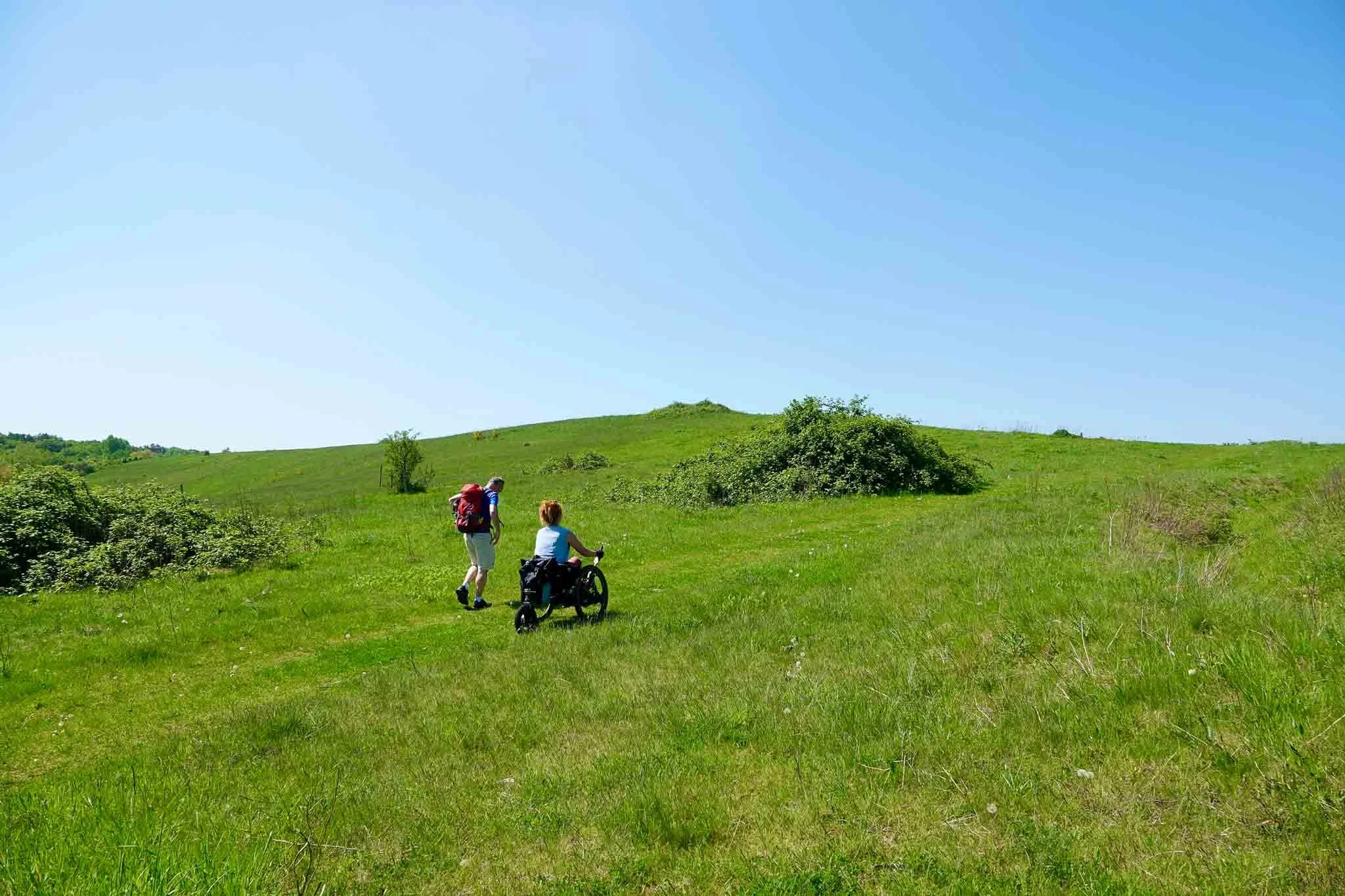 Twee mensen wandelen en één persoon zit in een rolstoel op een groene, open, heuvelachtige grasvlakte onder een heldere blauwe hemel.