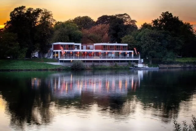 Een restaurant met grote ramen aan een rivier tijdens zonsondergang, omgeven door bomen.