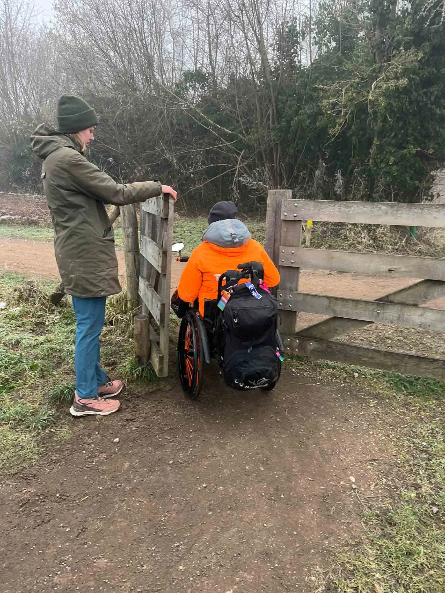 Een persoon in een rolstoel en een andere persoon staan bij een houten hek met een poort en kijken naar een open veld met bomen en struiken op de achtergrond.