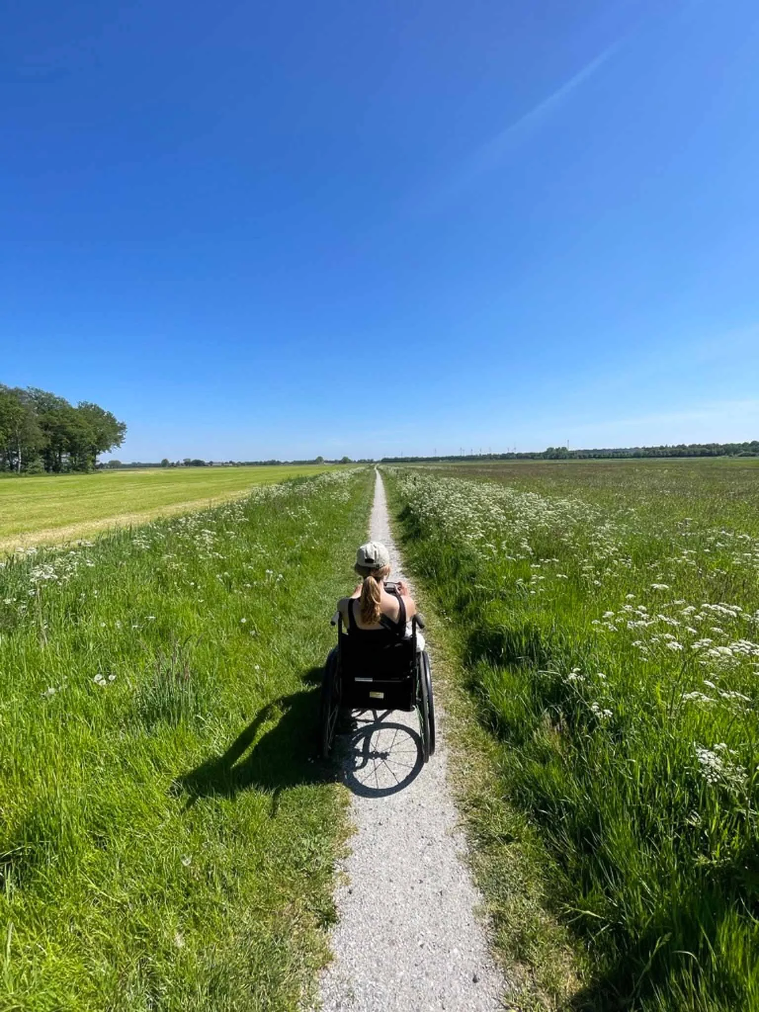 Persoon in een rolstoel fietst op een smal pad door een groen landschap met blauwe lucht.