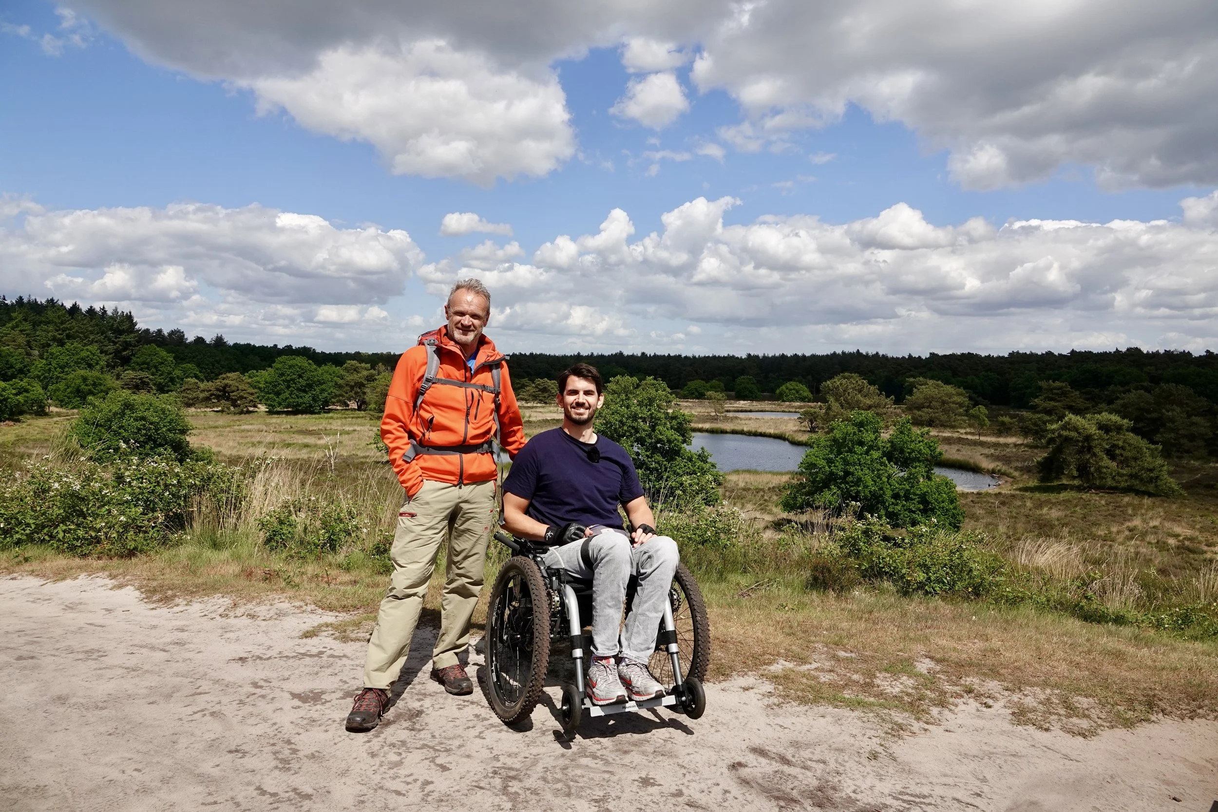 Twee mannen in een veld met een kleine meer op de achtergrond, een man in een rolstoel en een andere man die naast hem staat, beiden lachen en genieten van een buitenwandeling op een bewolkte dag.