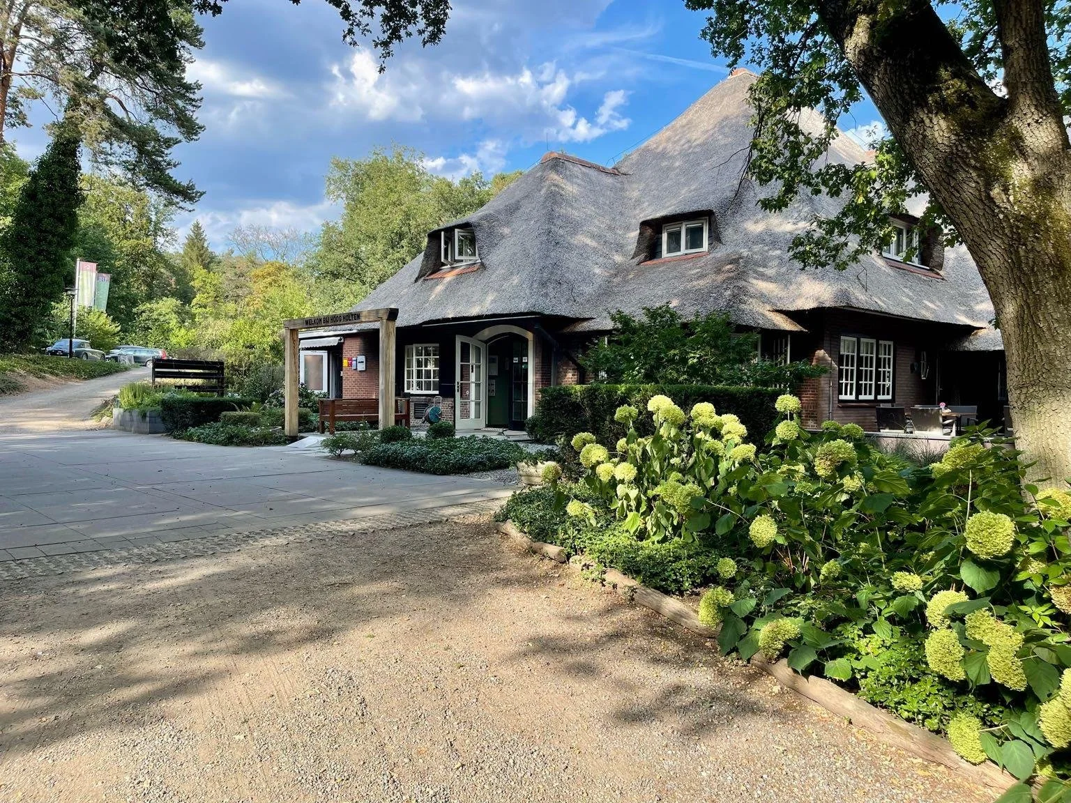 Toegankelijke accommodatie aan het Pieterpad. Een charmant huis met rieten dak, omringd door groene bomen en bloemen, langs een oprit met een bank en buitenmeubelen op een zonnige dag.