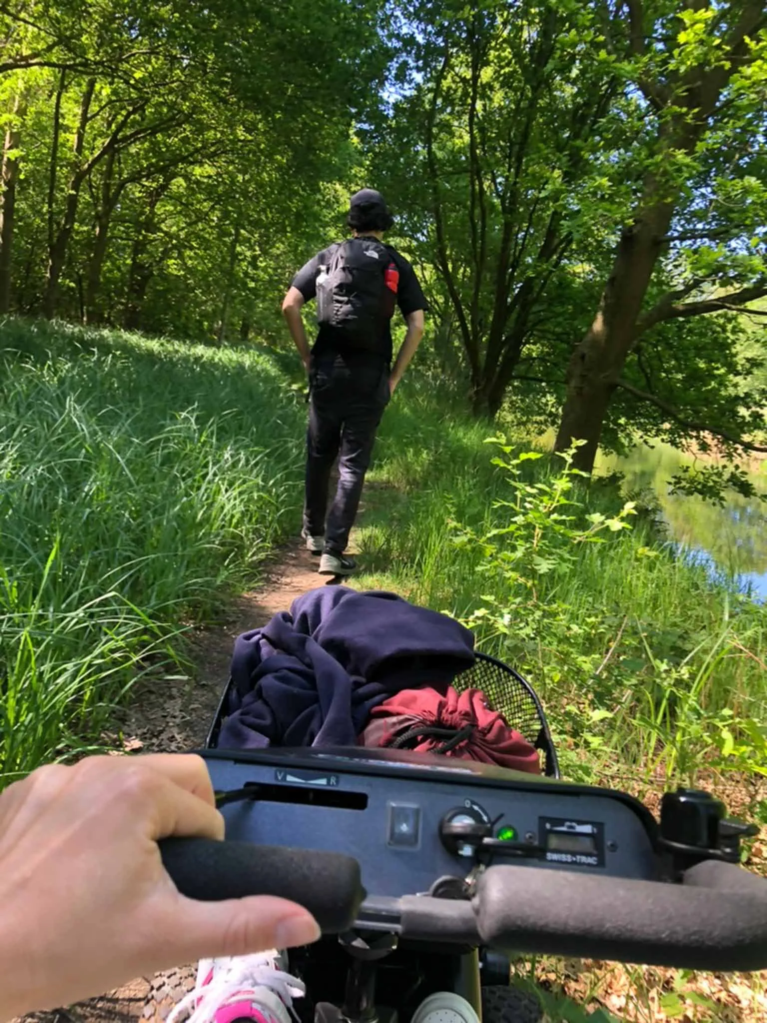 Een persoon rijdt op een elektrische driewielerfiets door een groen bos, terwijl een andere persoon verderop op een pad wandelt. De omgeving is bosrijk met veel groen en een rivier aan de rechterkant.