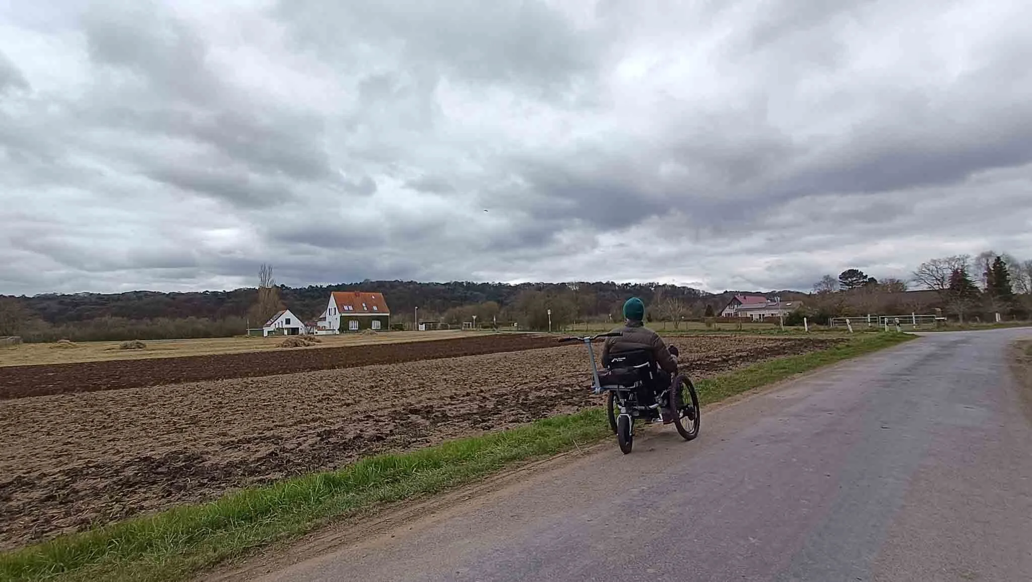 Persoon in een rolstoel rijdt langs een landweg met boerenvelden en huizen onder een bewolkte hemel.