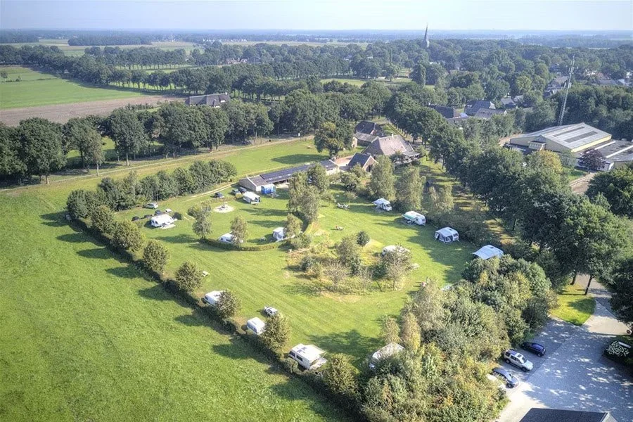 Luchtopname van een camping met tenten en caravans in een groene veldomgeving, omgeven door bomen en landbouwgebieden, met enkele gebouwen op de achtergrond.
