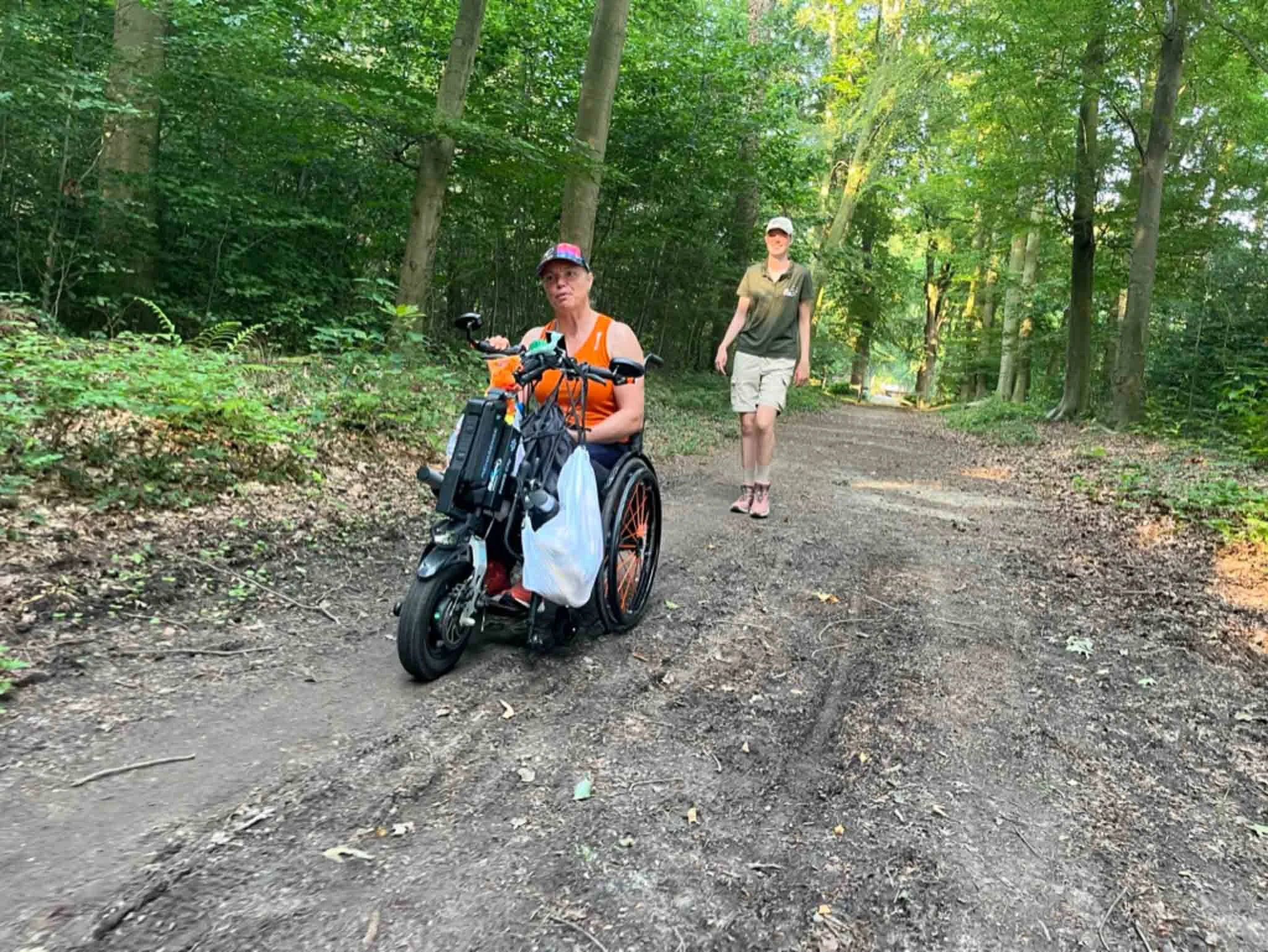 Twee vrouwen wandelen in een bos; een vrouw in een elektrische rolstoel met een oranje top en een vrouw in een groene blouse en beige korte broek.