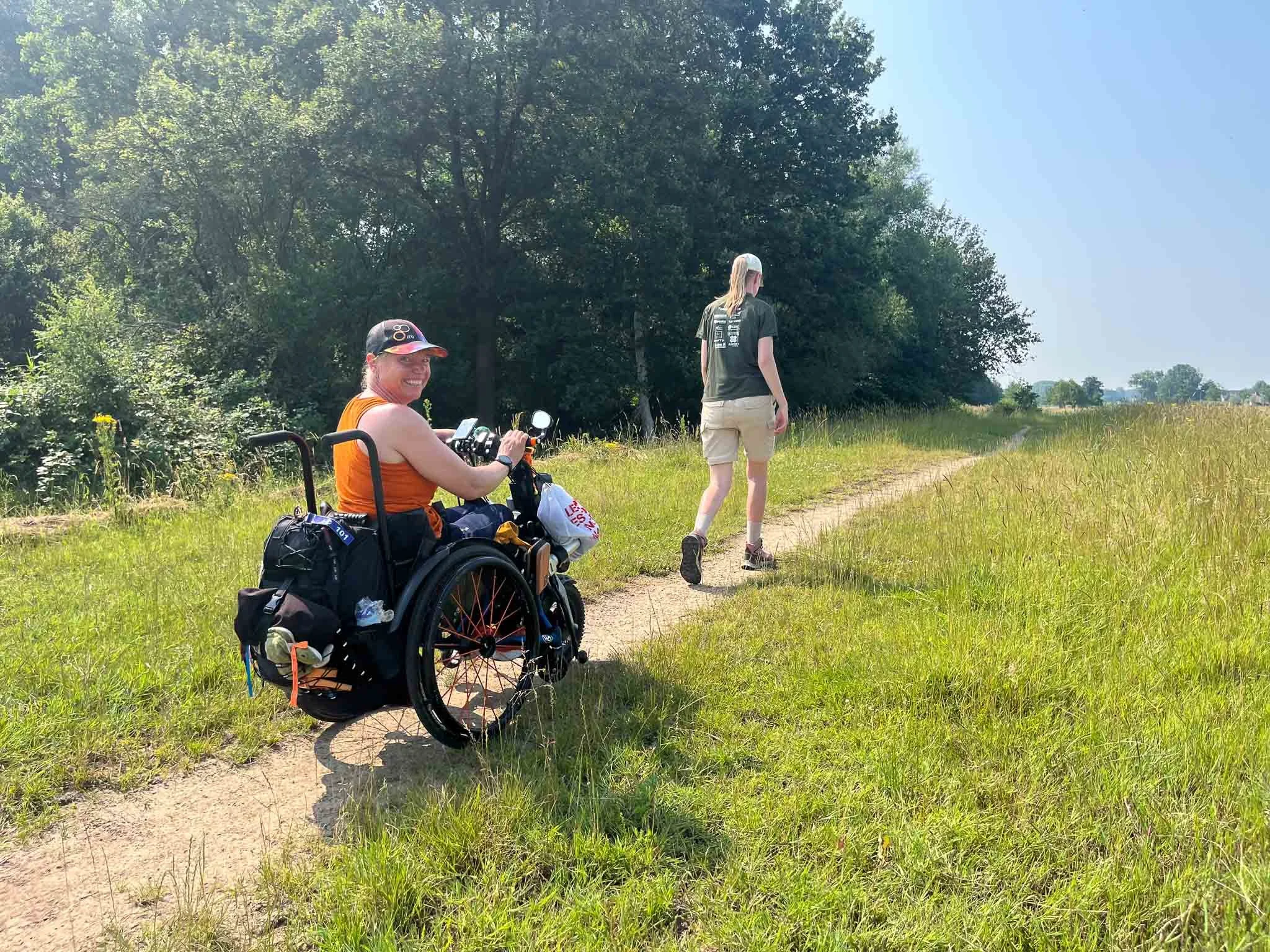 Twee mensen wandelen op een smal pad in een open veld met bomen op de achtergrond. Eén persoon zit in een rolstoel en kijkt lachend naar de camera, terwijl de ander verder weg wandelt.