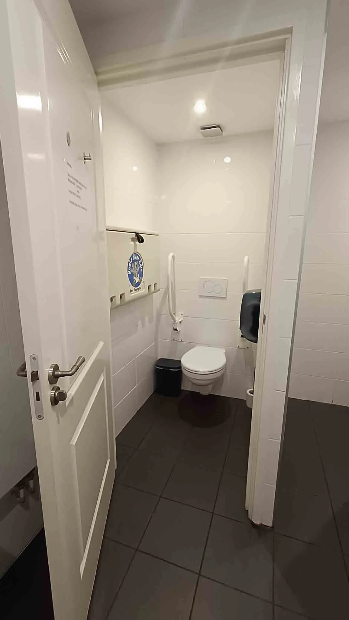 Klantentoilet met witte wc, zwart prullenbak, donkere vloertegels, witte muren en plafond, automatische plafondverlichting, douchestang en handgreep