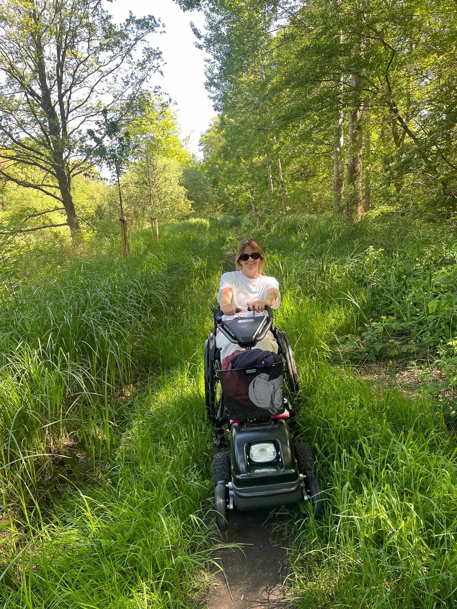 Vrouw in een rolstoel op een smalle bosweg, omringd door groen gras en bomen, genietend van de natuur.