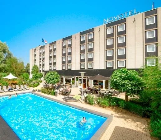 Het hotelgebouw 'Novotel' met zwembad en ligstoelen op de terras. Er is een persoon in het zwembad en tuinmeubilair onder bomen.